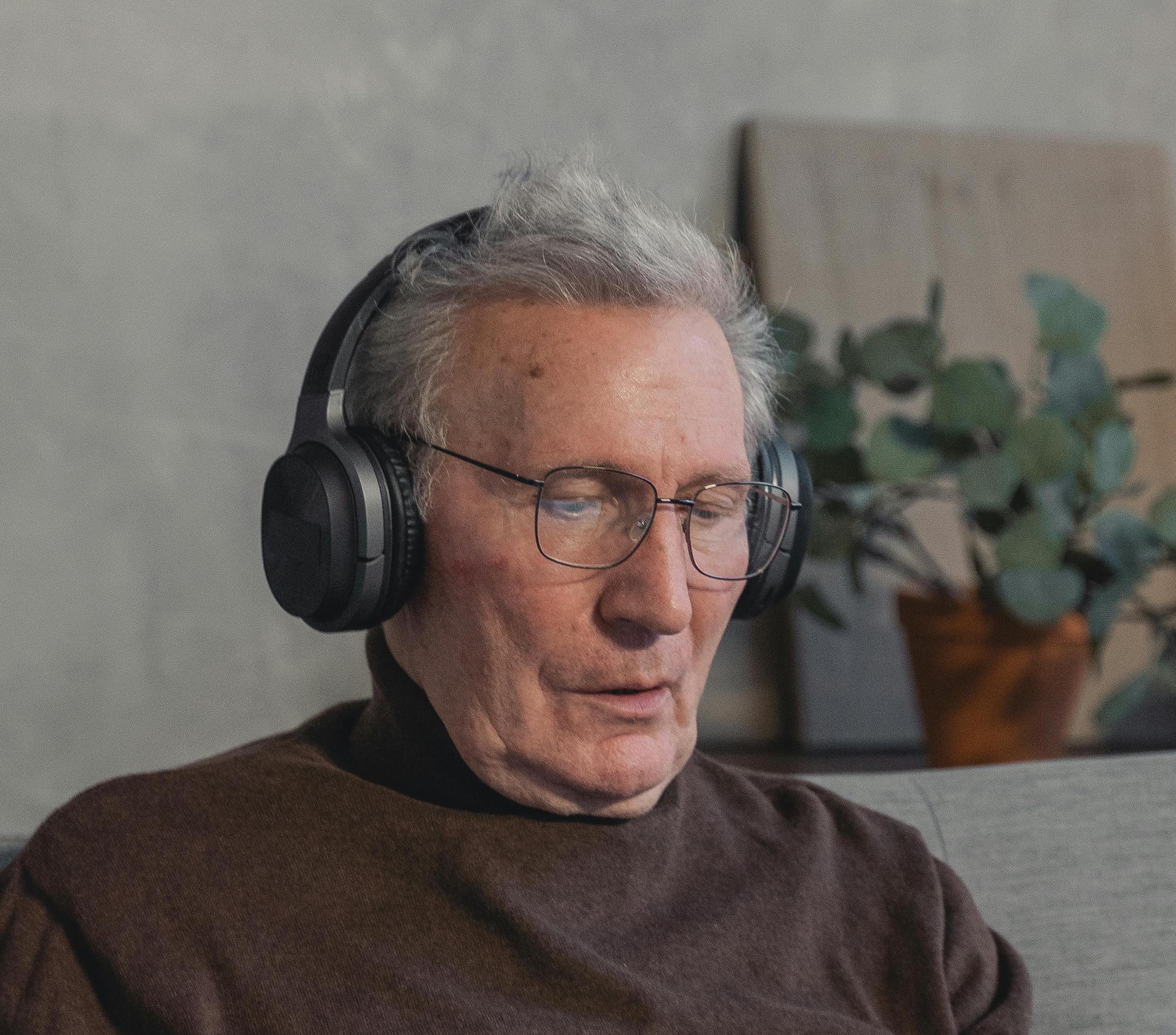 An elderly man with gray hair and glasses is wearing large black over-ear headphones and is looking at a device or screen.
