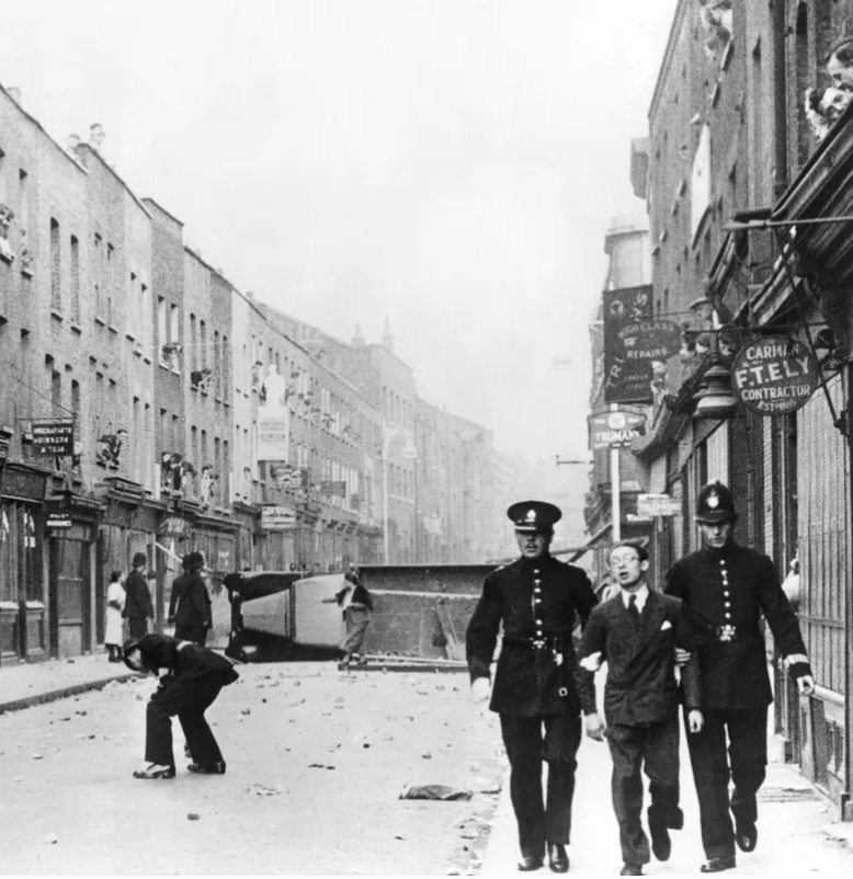 A black and white photo of a city street with police officers escorting a young person, with buildings, storefronts, and pedestrians in the background.