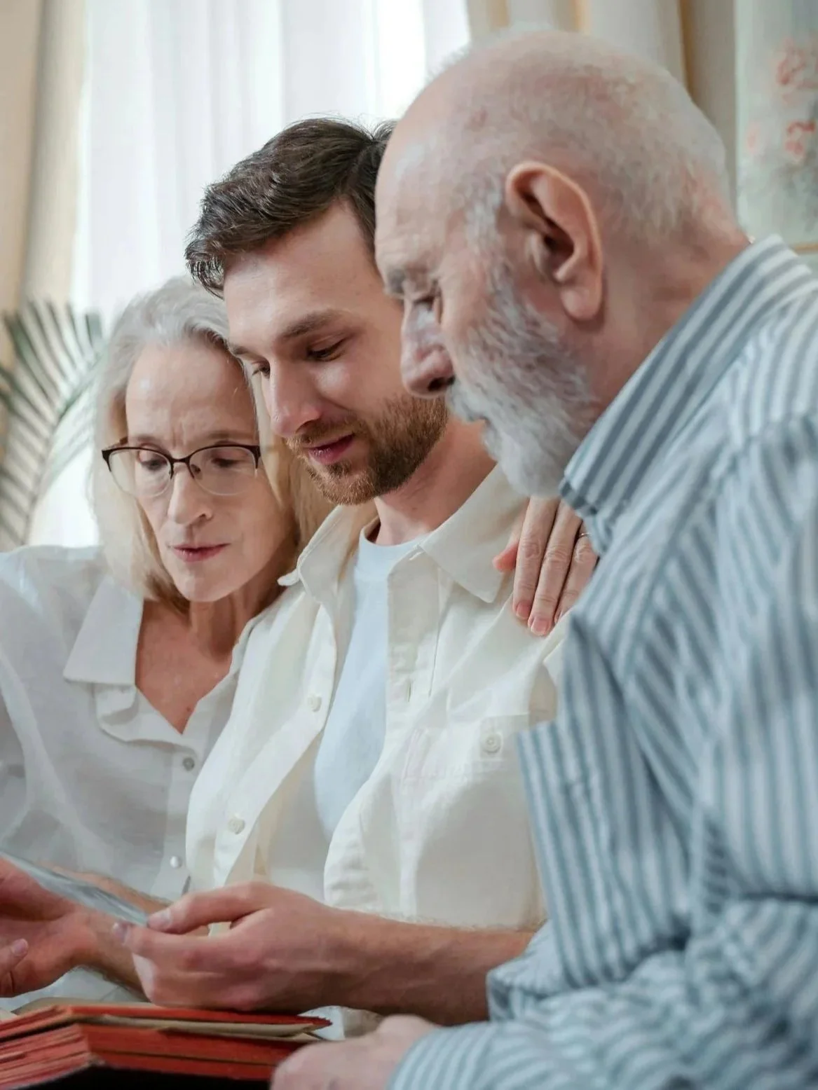 Three people, an elderly woman, a young man, and an elderly man, are closely looking at a photo album together indoors.
