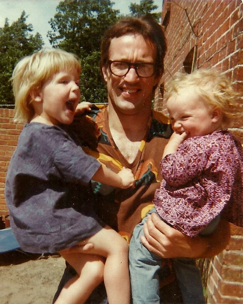 A man with glasses holding two young girls outside near a brick wall, all smiling and laughing on a sunny day.