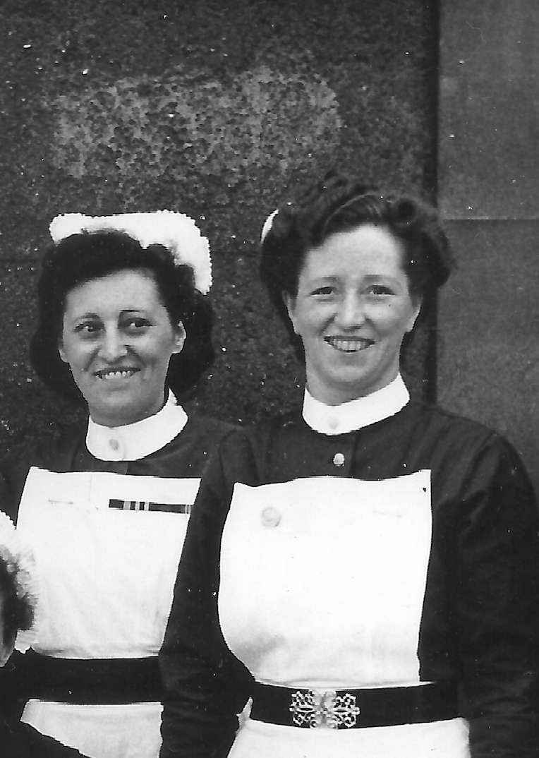 Two women dressed as nurses, standing outside, smiling at the camera. Both have white collars and aprons, one with a flower-shaped belt buckle, and one with a white headpiece.