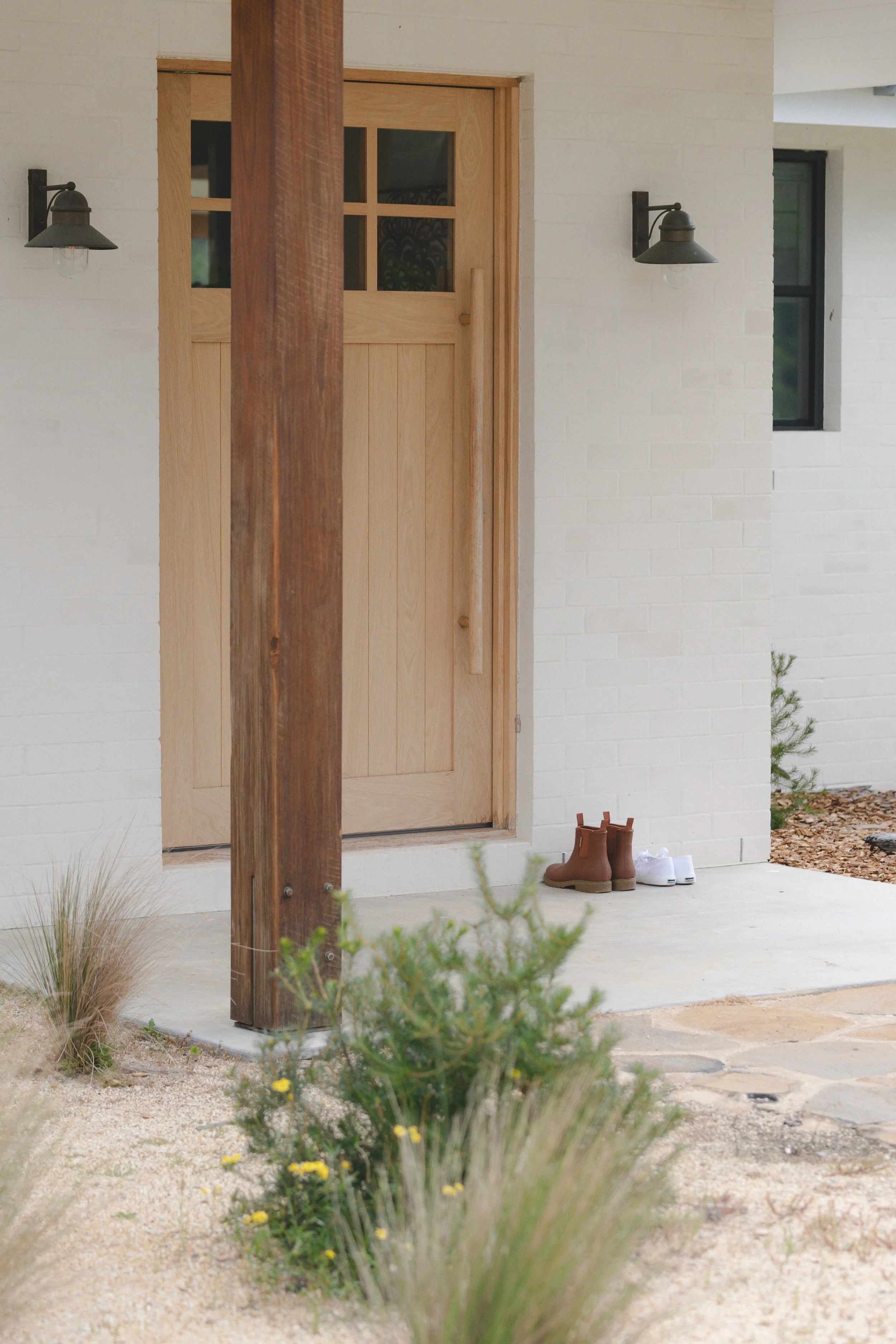 A front door of a house with two pairs of shoes, one brown boots and one white sneakers, on the porch. Two exterior wall-mounted lights are on each side of the door. The house has white brick walls, a black window to the right, and some plants and dr