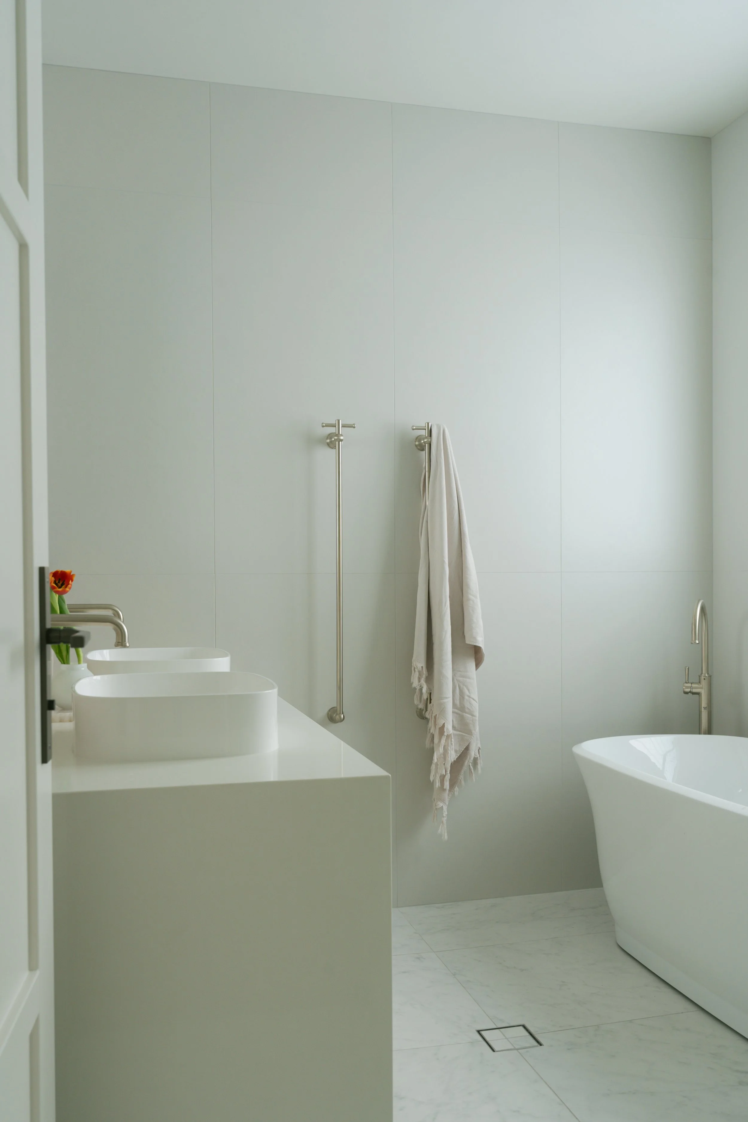 A modern, minimalist bathroom with white walls, a white freestanding bathtub, a white sink with a bouquet of flowers, and a towel hanging on a wall-mounted bar.