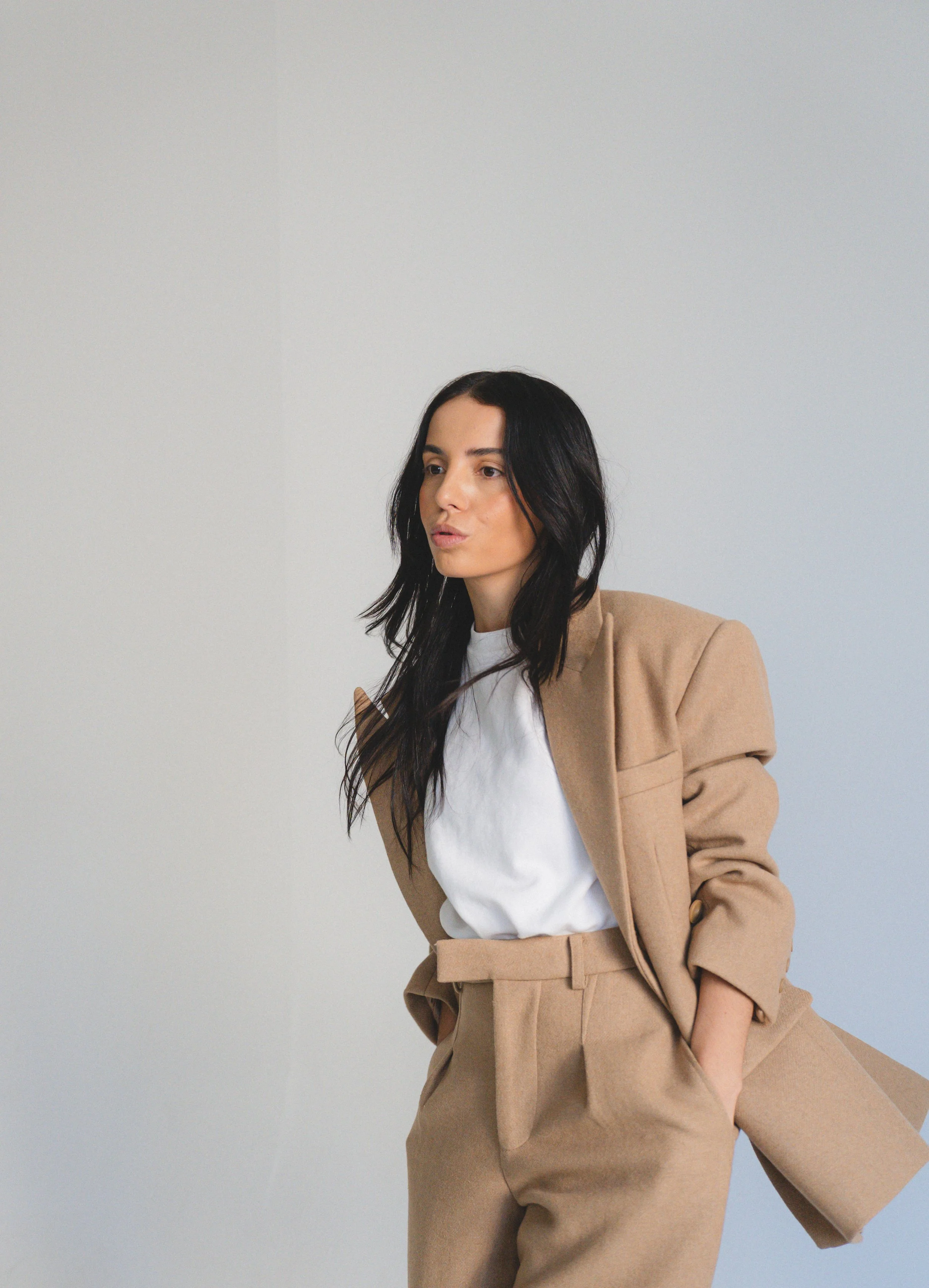 A woman with long dark hair wearing a beige blazer and matching pants, with a white shirt underneath, standing against a plain light gray background.