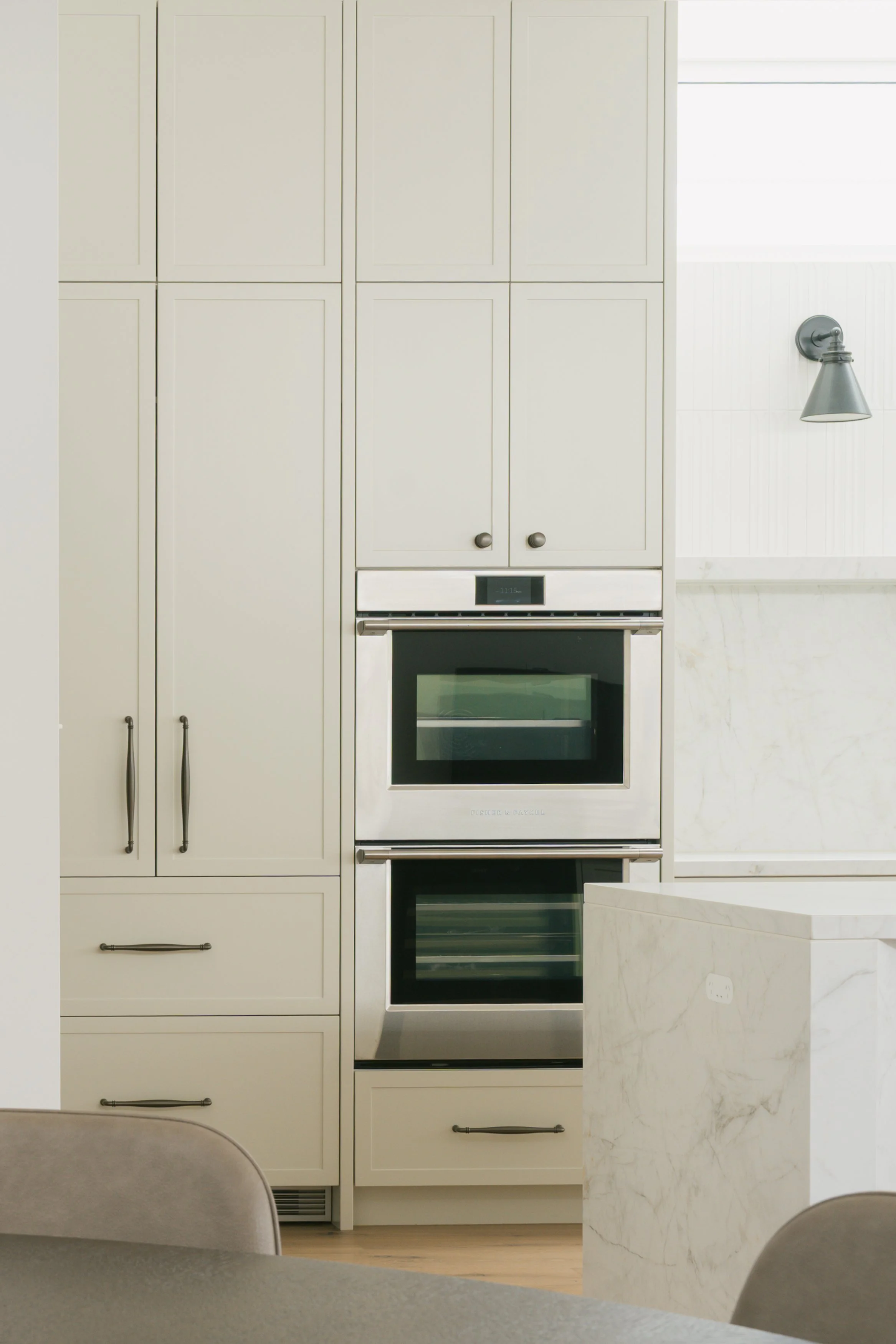 A modern kitchen with white cabinets and a built-in oven, a gray wall-mounted light, and a marble kitchen island in the foreground.
