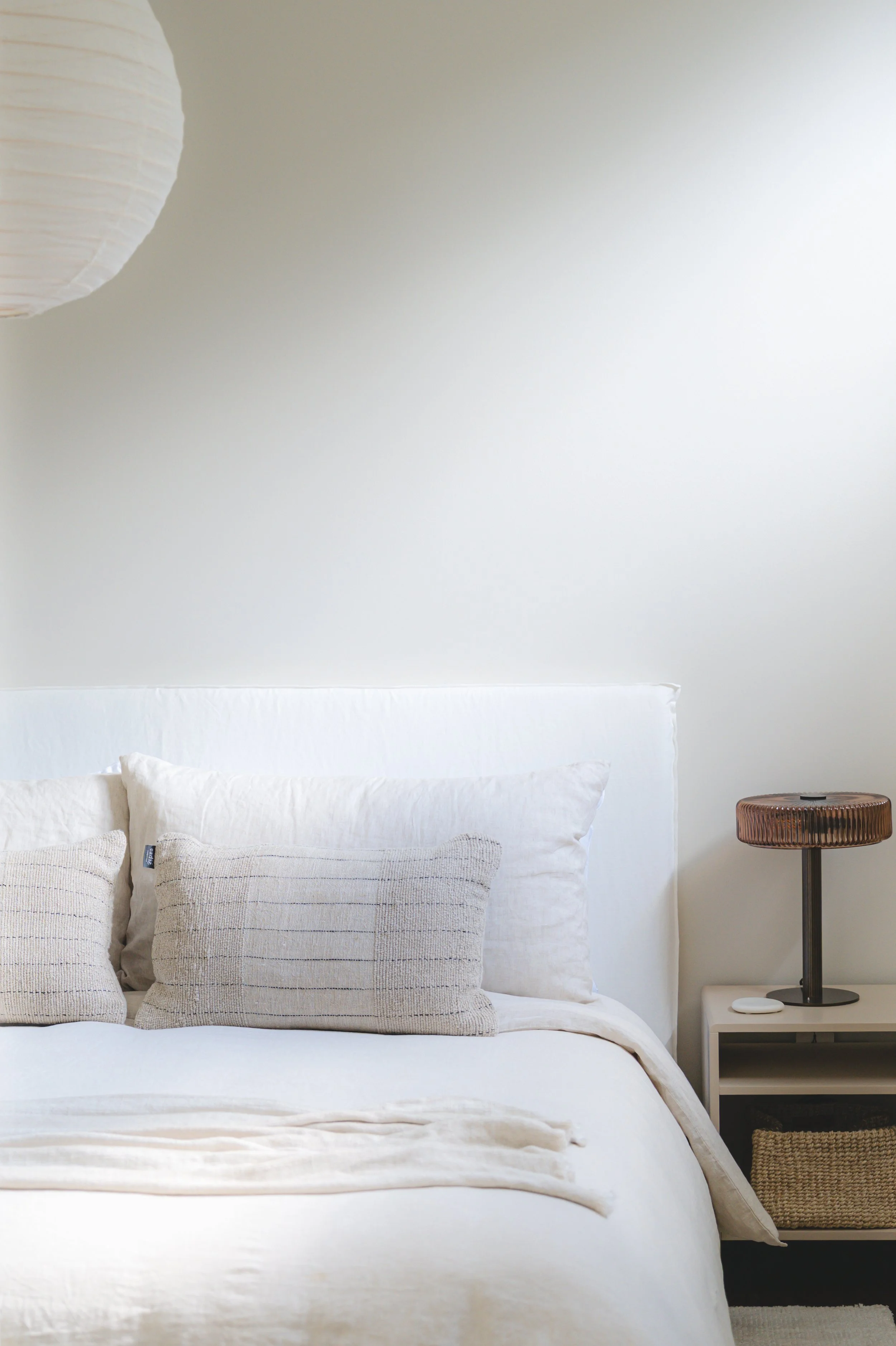 A neatly made bed with white bedding and textured beige pillows, a nightstand with a decorative table lamp, on a minimalist bedroom wall with a white and cream color scheme.