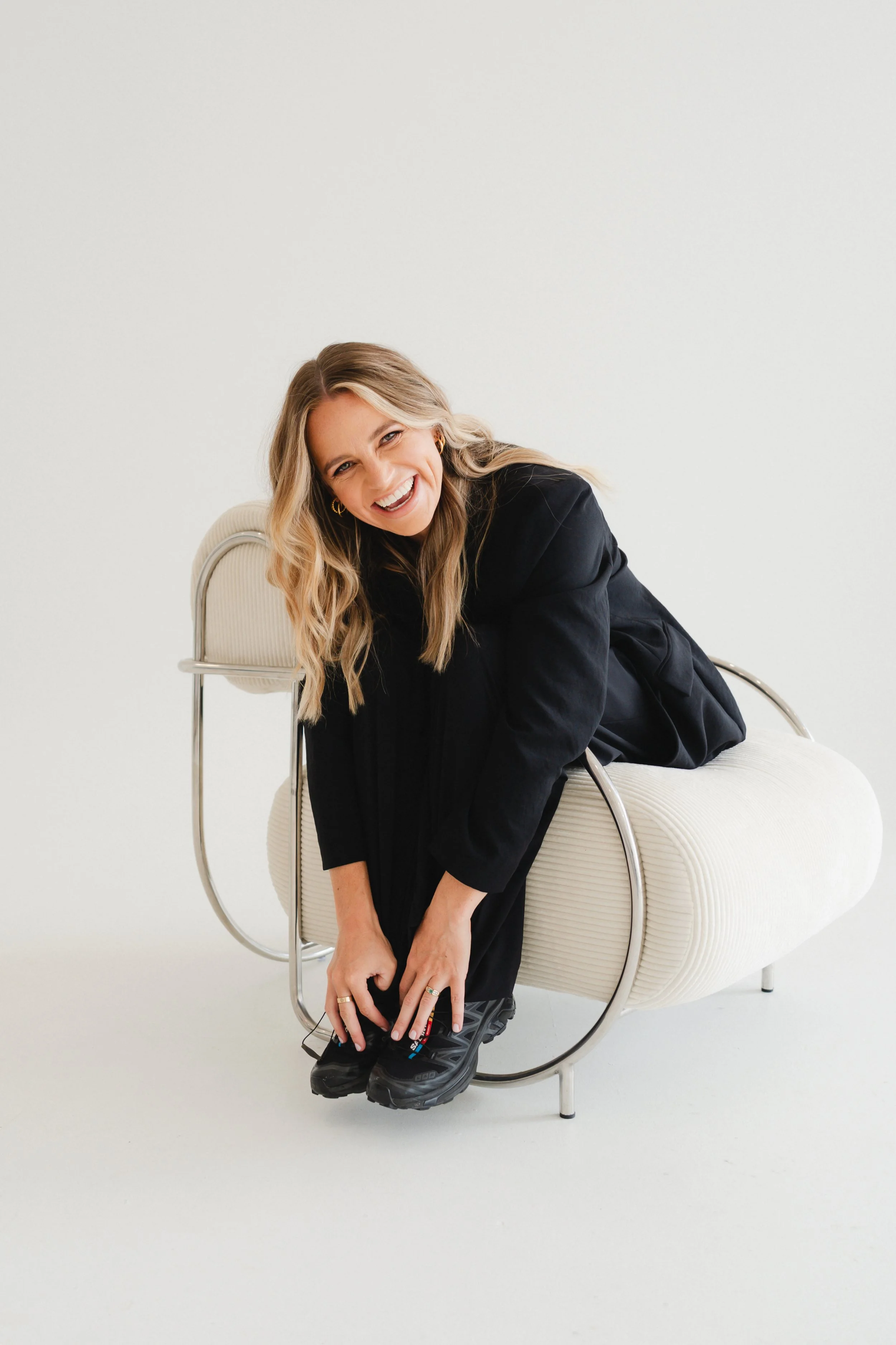 A woman with long blonde hair, smiling, wearing a black oversized shirt, sitting on a modern beige chair with metal armrests, against a plain white background.