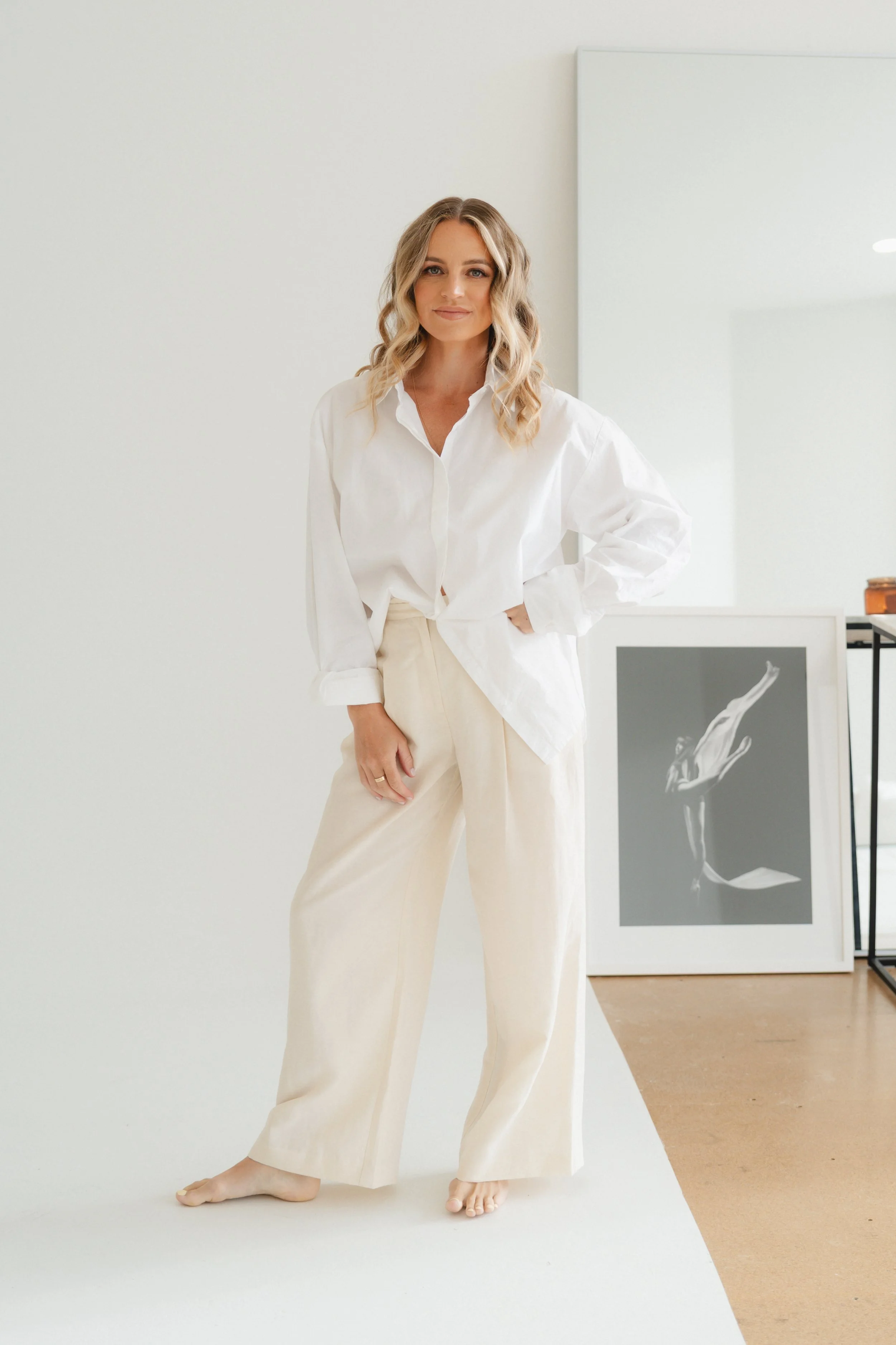 A woman with blonde wavy hair wearing a white shirt and beige pants standing in front of a minimalistic white wall, with some artwork on the wall to her right.