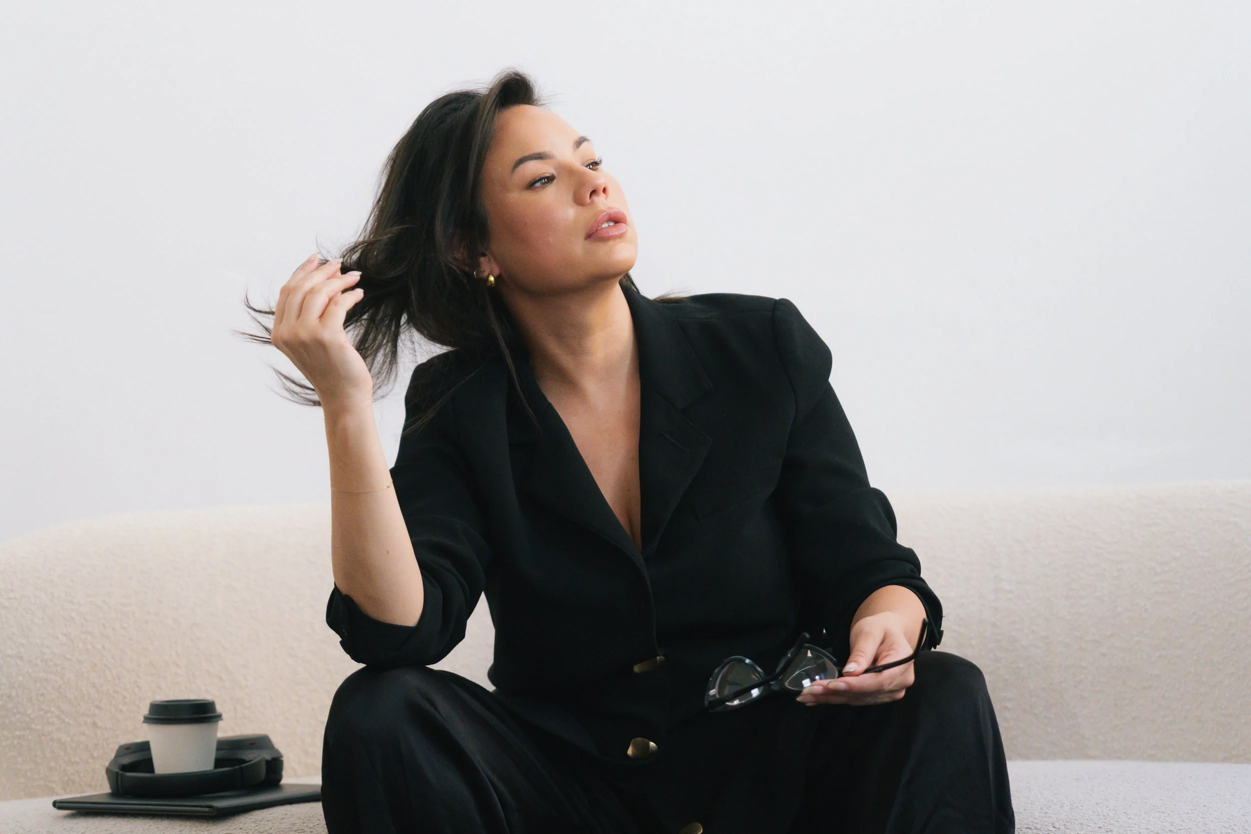 A woman sitting on a beige couch holding eyeglasses in her right hand, with a coffee cup and a pair of glasses on the table in front of her, against a plain white wall background.