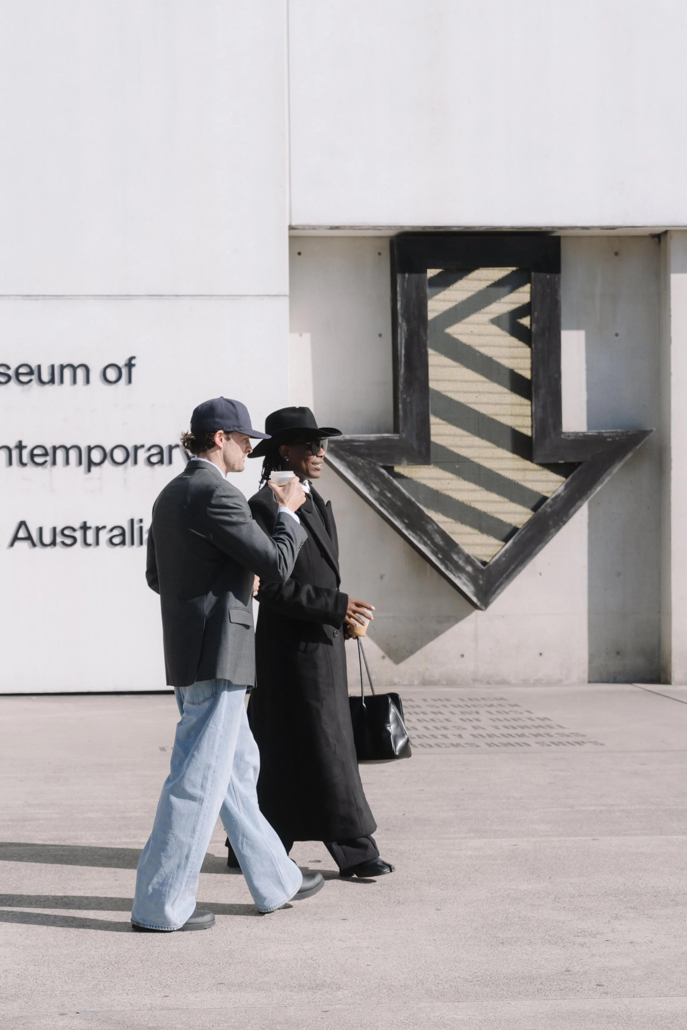 Two people walking outside a museum of contemporary art in Australia, with large geometric artwork on the wall behind them.