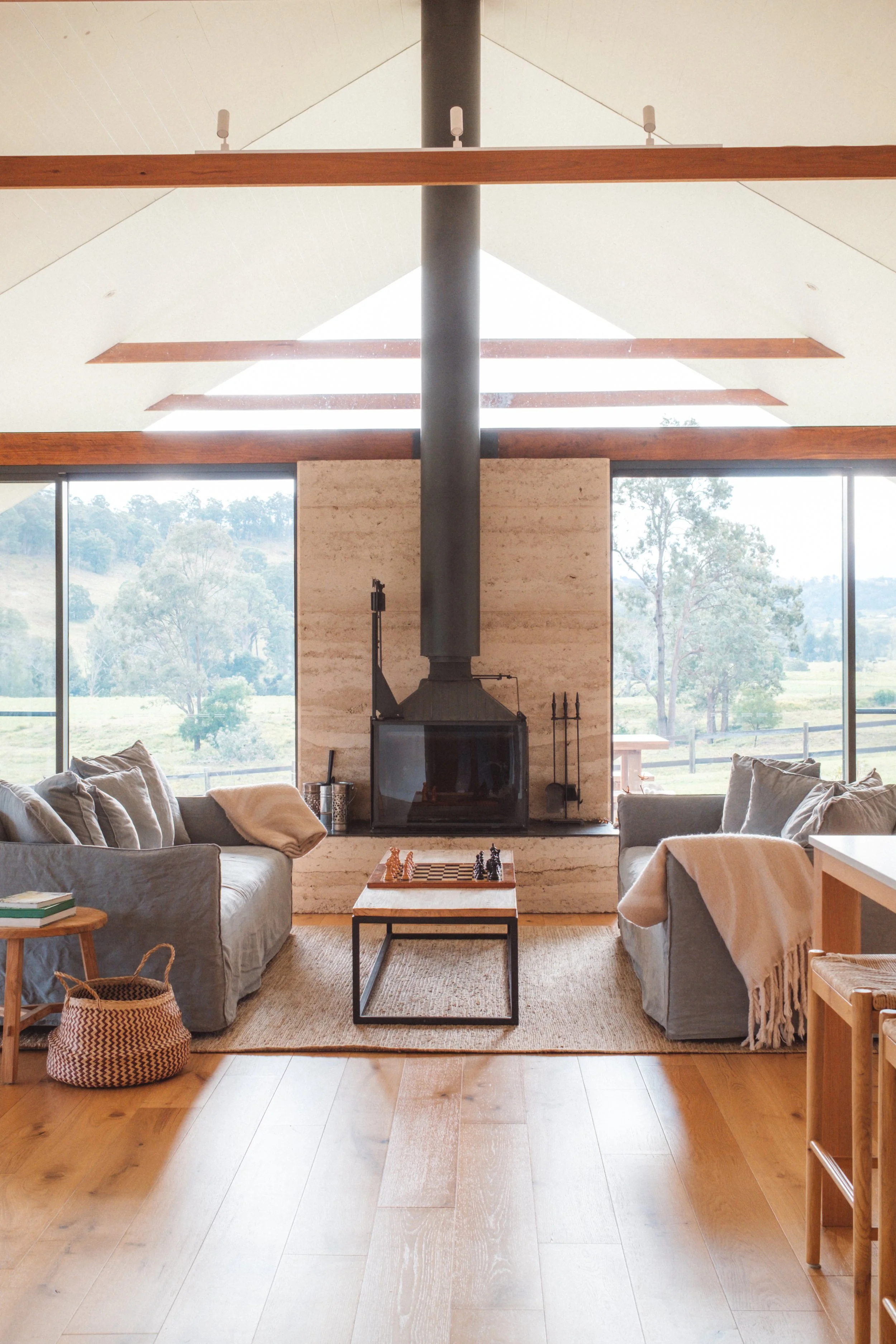 Living room with two sofas, a coffee table with chess, a fireplace with a large pipe, large windows showing outdoor scenery, and a wooden floor.
