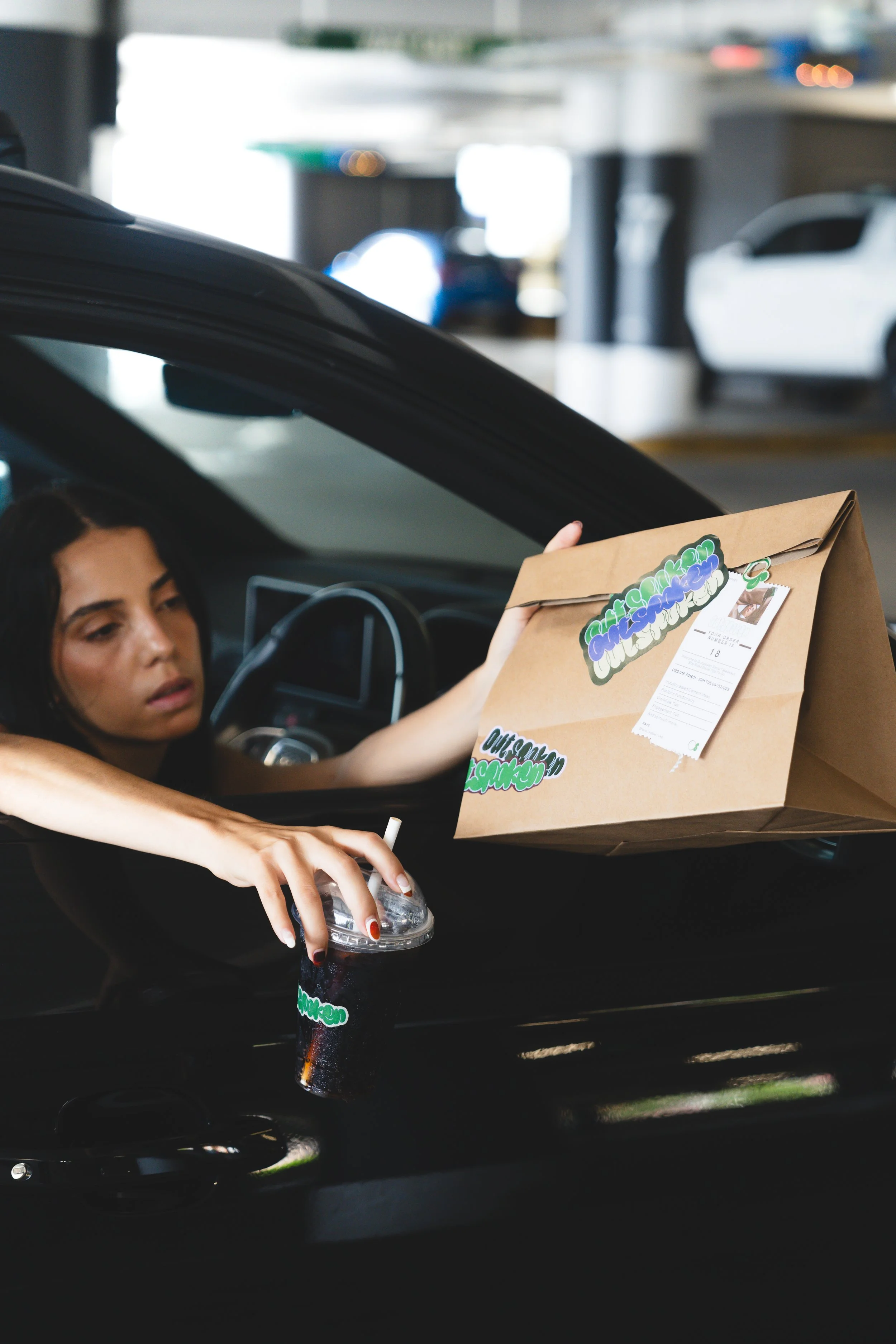 A woman with dark hair sitting in a car, holding a brown paper bag with colorful stickers and a boarding pass, and reaching for a drink from a drive-thru window.