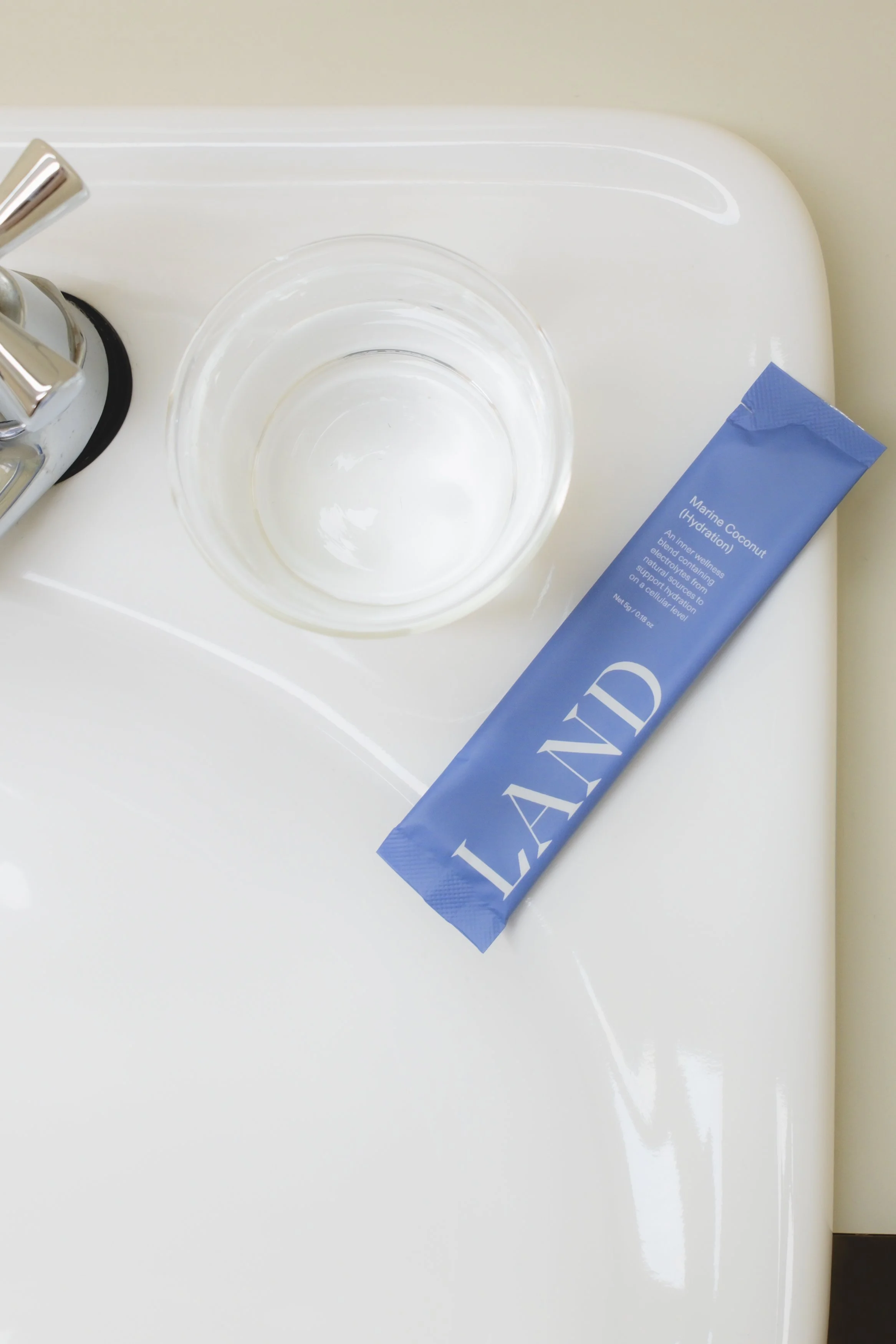 A white bathroom sink with a chrome faucet, a clear glass bowl, and a tube of skincare product labeled 'Marine Coconut Hydration' placed on the sink's surface.