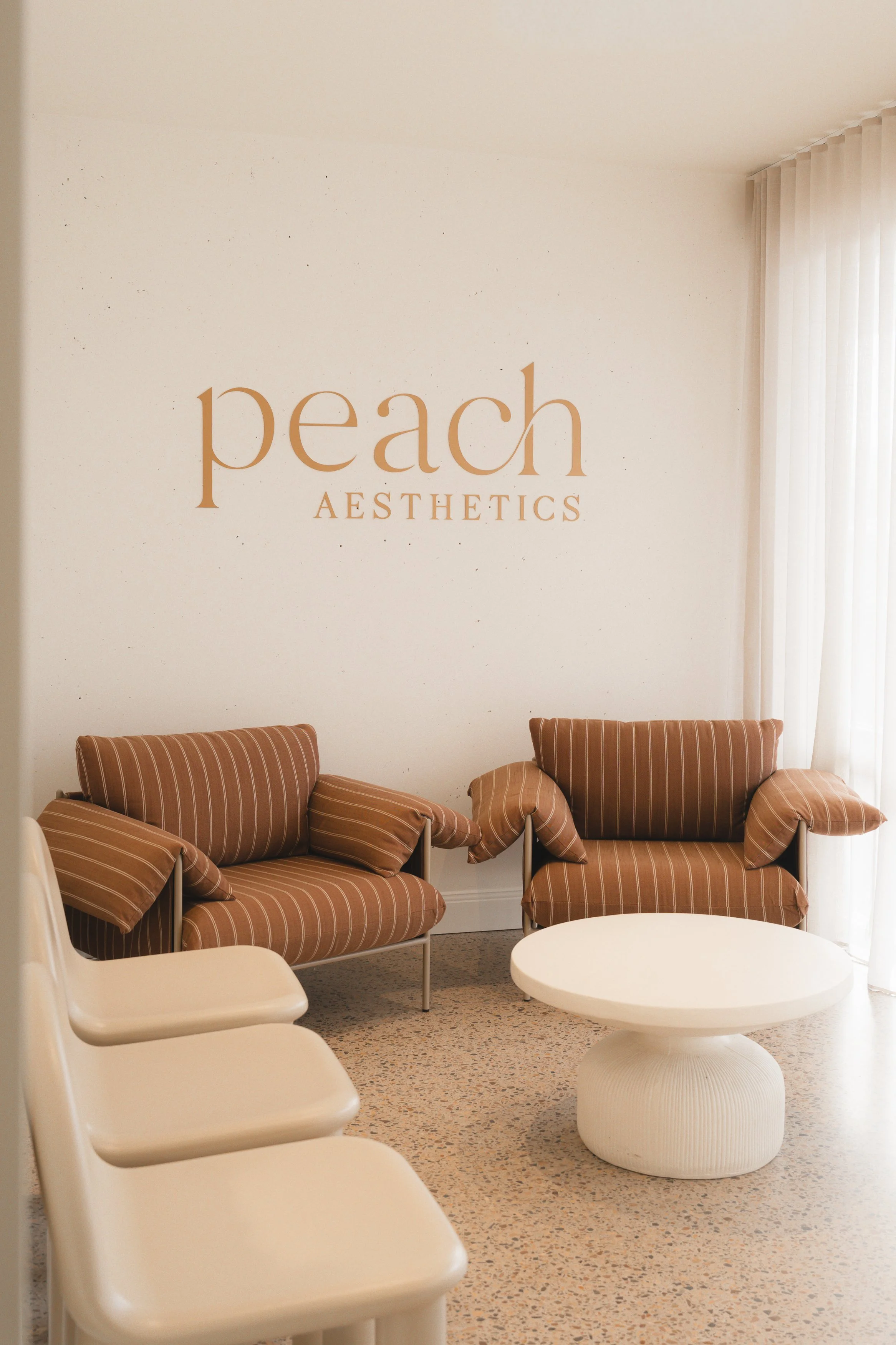 A waiting area with three striped armchairs, a white round coffee table, and a white chair. The wall behind has a sign that says 'peach AESTHETICS'. There is a window with vertical blinds on the right.