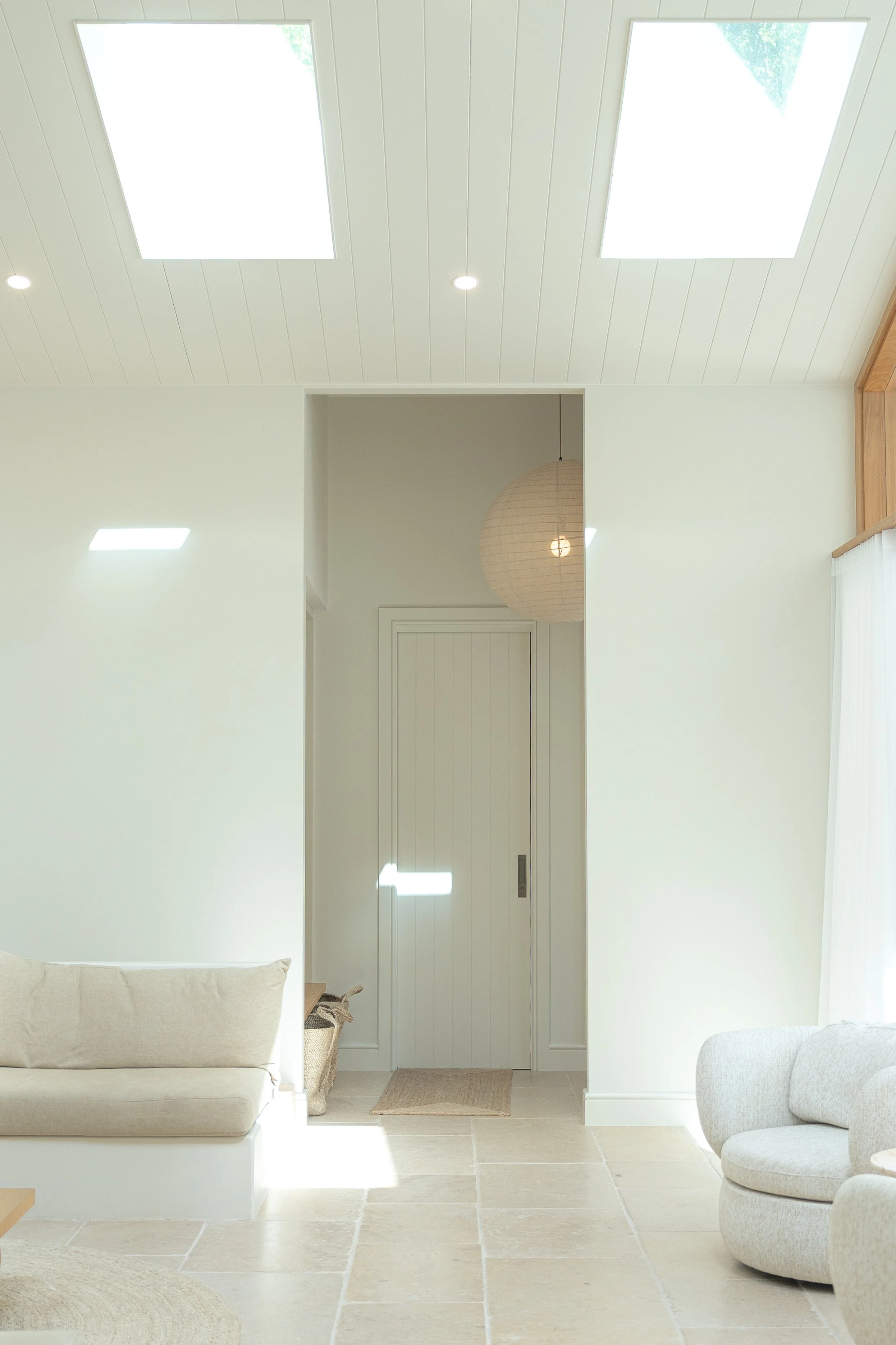 Bright, minimalistic living room with natural light streaming through skylights. Off-white walls and large beige tile flooring. Comfortable beige sofa and a gray upholstered chair are visible, with neutral decor and a round paper lantern and woven ma