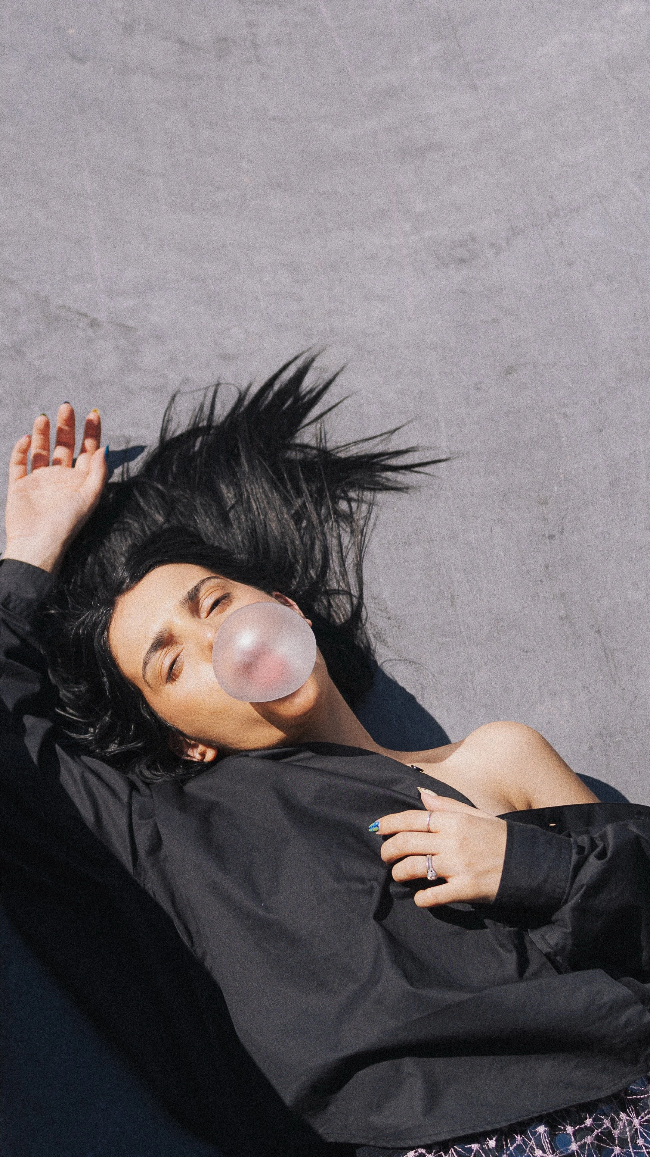 A woman with long black hair lying on the ground, blowing a bubble gum bubble, wearing a black jacket and patterned pants.