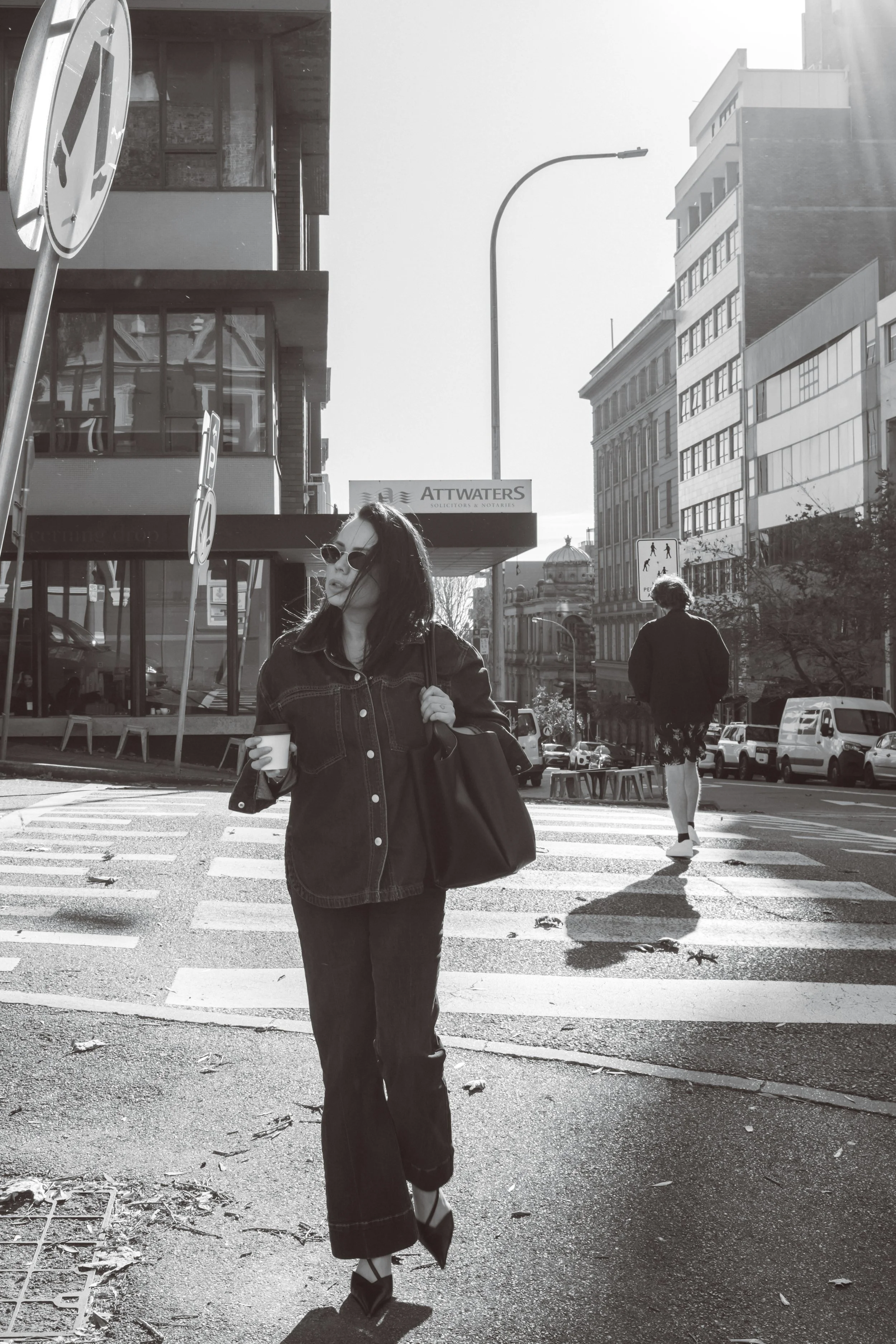 A woman with dark hair wearing sunglasses, a denim jacket, and dark pants, holding a cup in one hand and a large bag in the other, standing at a crosswalk in an urban area with buildings and cars, and a man walking ahead in the background.