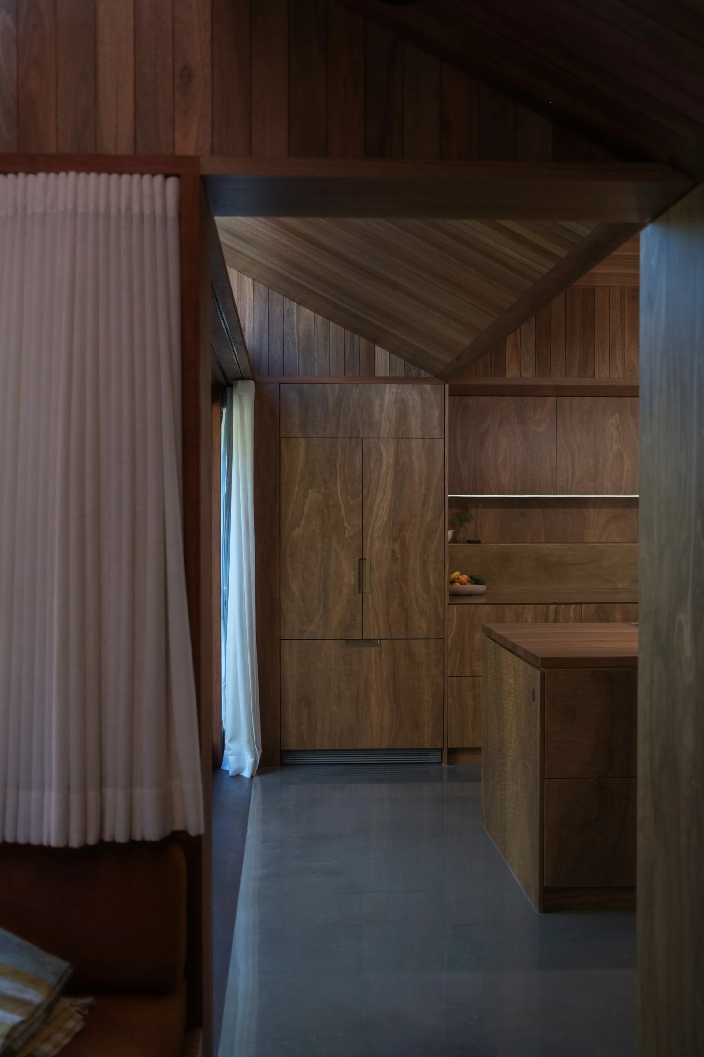 A wooden interior space with wood-paneled walls, ceiling, and cabinetry. A white curtain partially covers a window, and a small bowl of fruit is on a shelf.