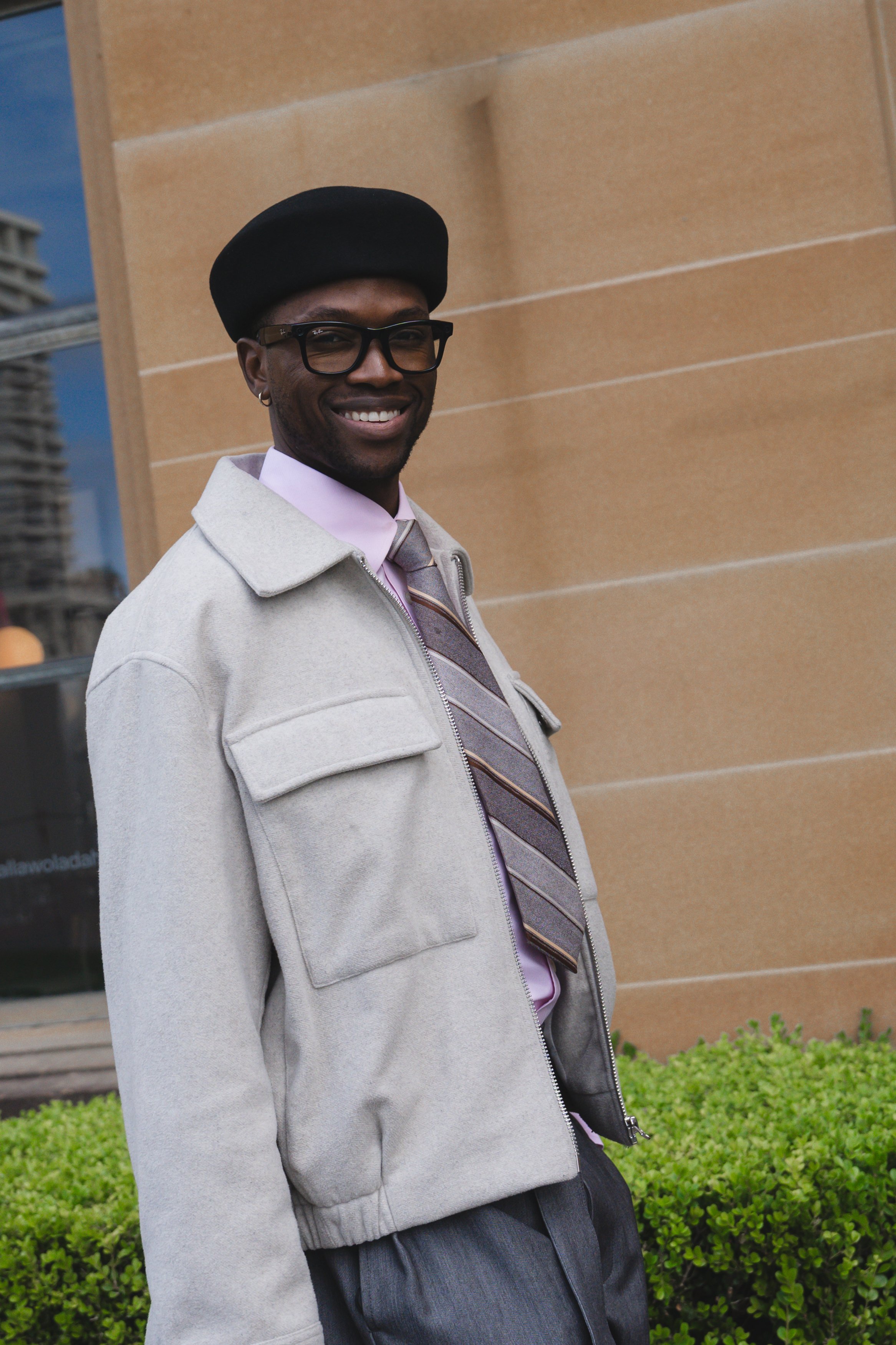 A man with dark skin, wearing glasses, a black beret, a light gray jacket, a pink shirt, and a striped tie, smiling and standing outdoors against a brick wall with greenery below.
