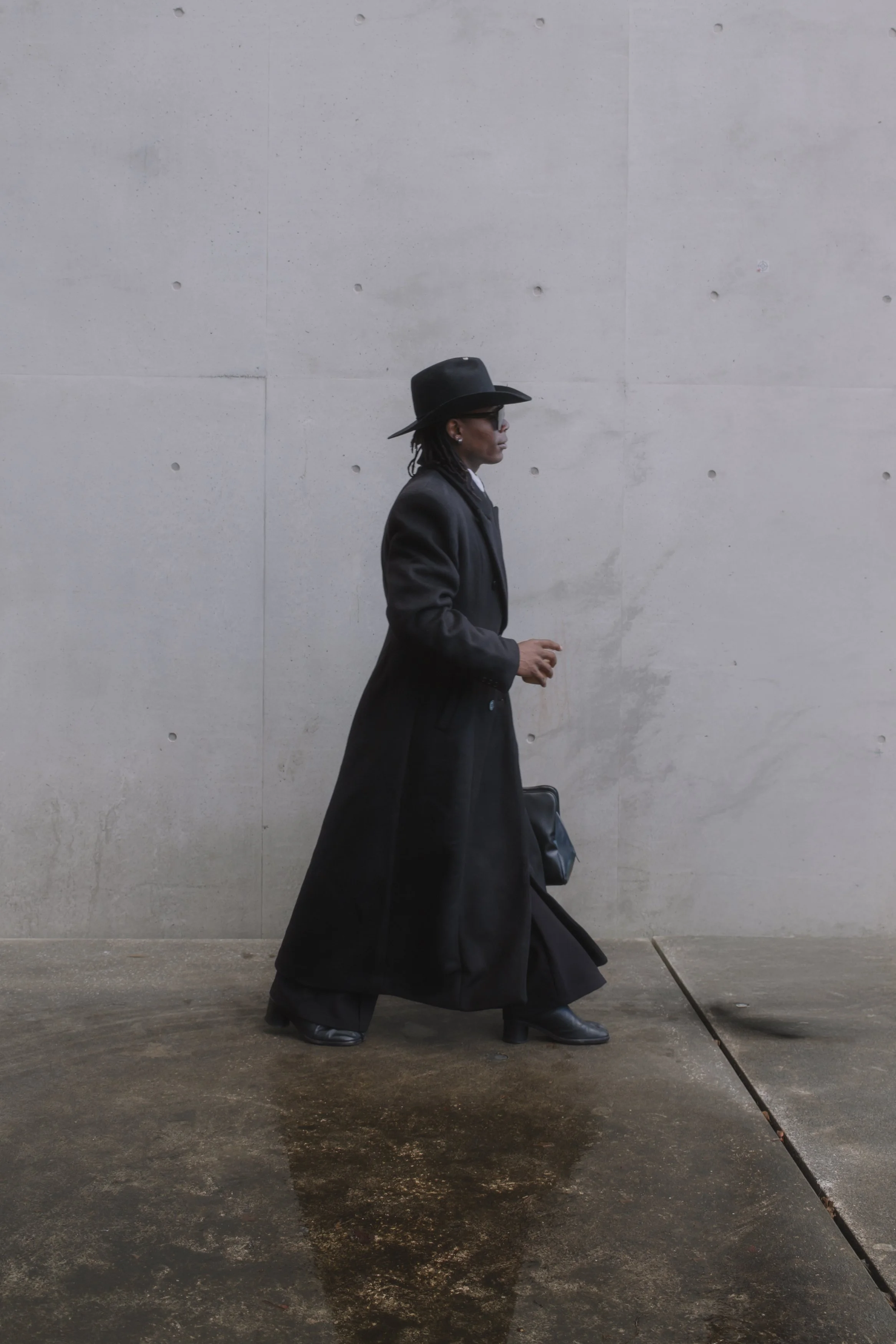 A person wearing a black hat, glasses, a long black coat, and black boots, walking against a plain light gray wall on a concrete sidewalk.