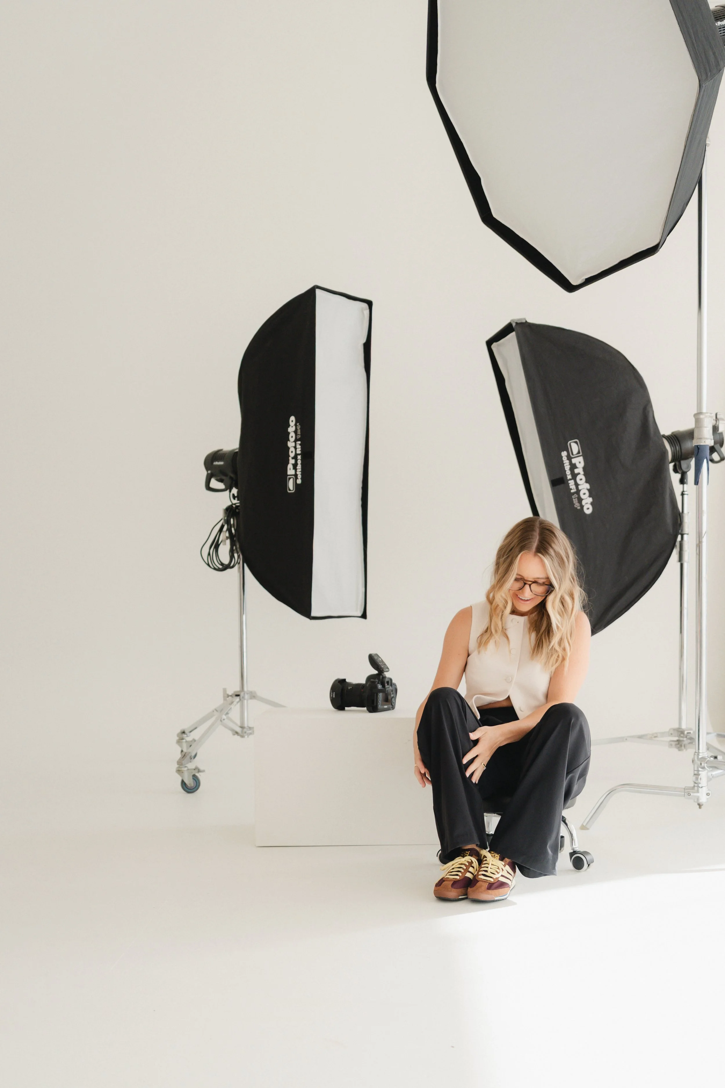 A smiling young woman with blonde hair and glasses sitting on the floor in a professional photography studio, surrounded by large softbox lights, with a professional camera on a stand nearby.