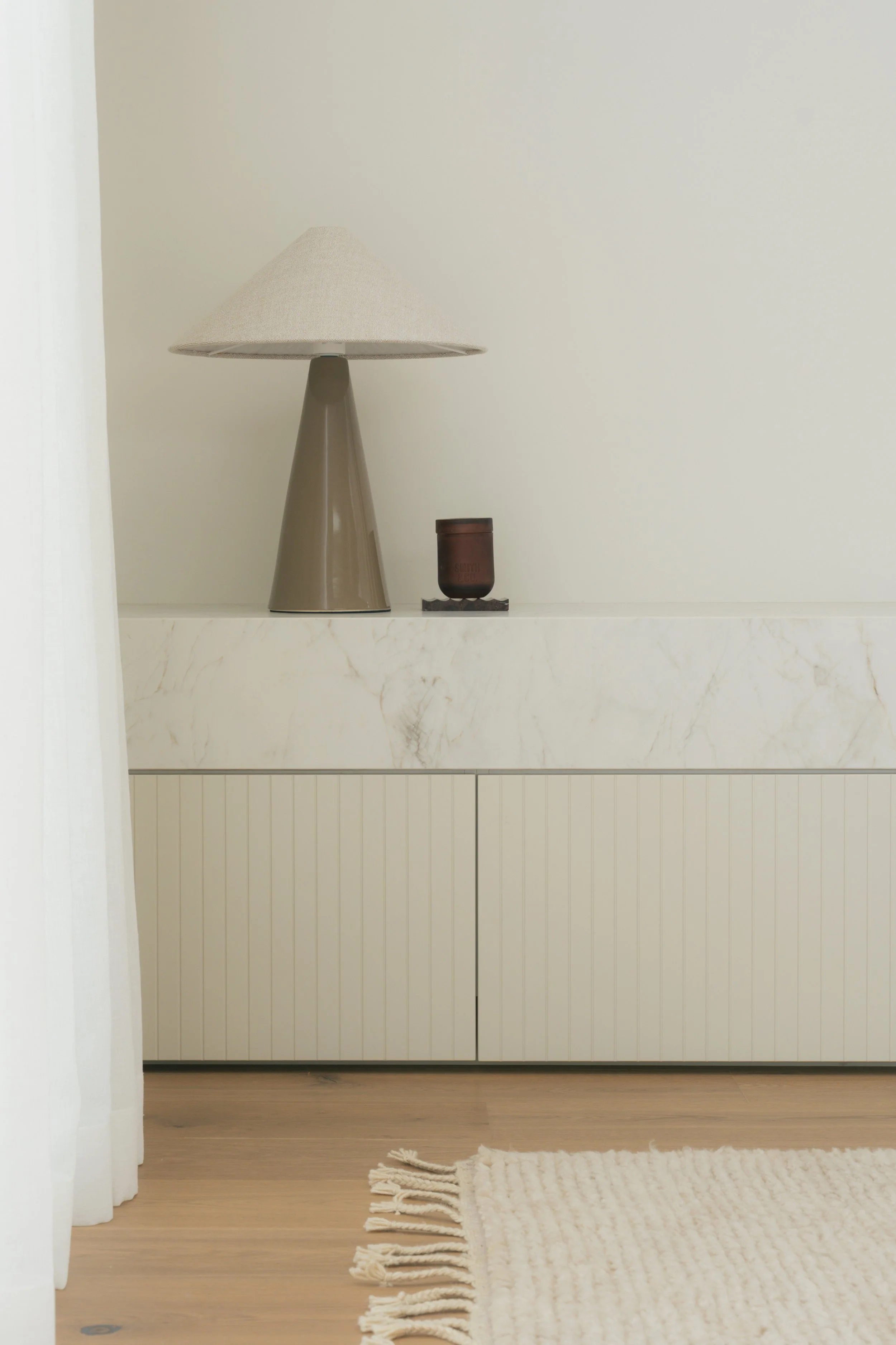 A minimalist interior sideboard with a beige table lamp and a small dark brown vase on top, a white textured rug on light wooden floor, and a plain white wall background.