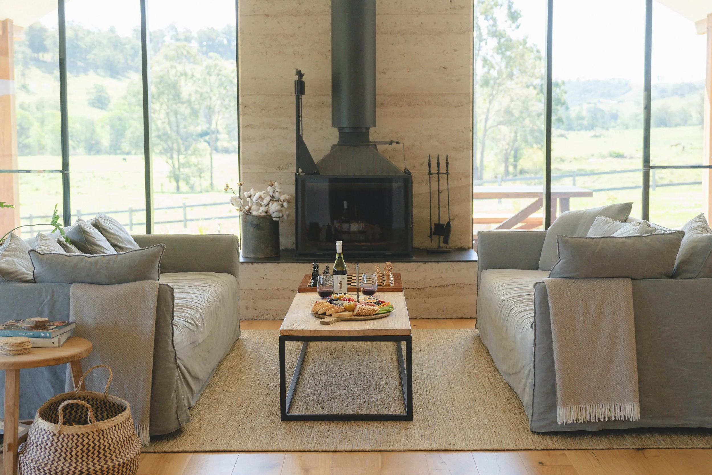 Living room with large windows showing a green outdoor landscape, two gray sofas facing each other, a wooden coffee table with wine, cheese, and snacks, a potted cotton plant, and a fireplace with a chimney.