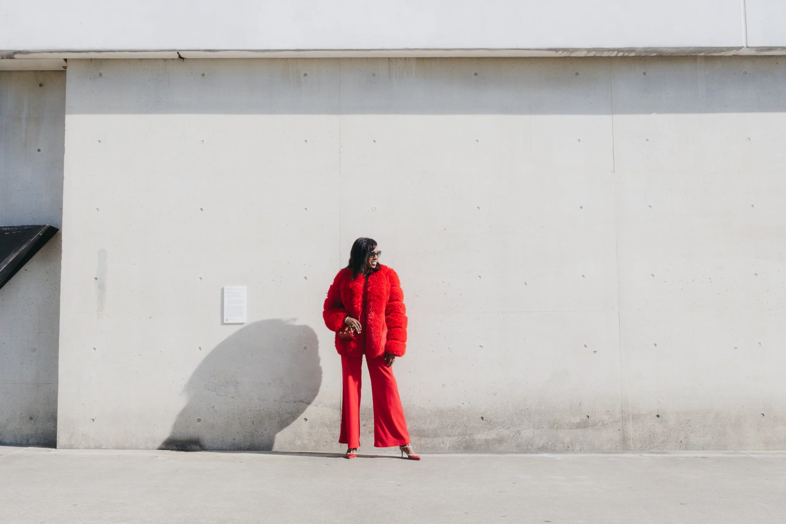 A woman dressed in a bright red furry coat and red pants is standing against a plain concrete wall. She is smiling, wearing sunglasses, and appears to be enjoying a sunny day.