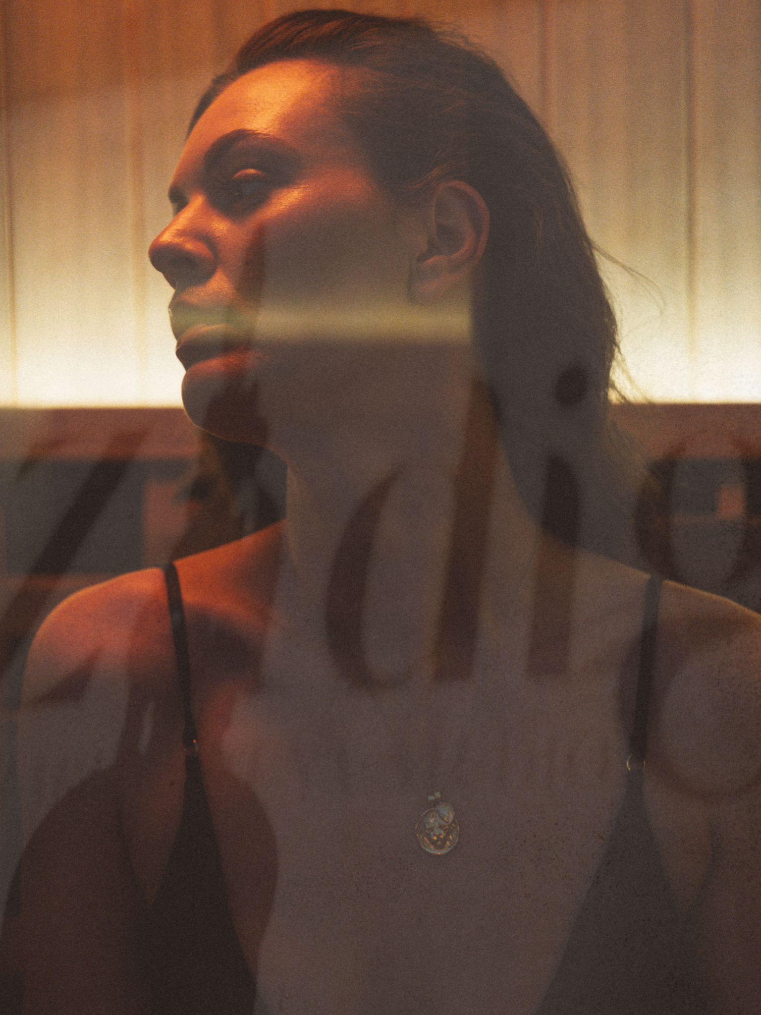Close-up of a woman with blonde hair wearing a necklace with a round pendant, photographed through a reflective surface with warm lighting.
