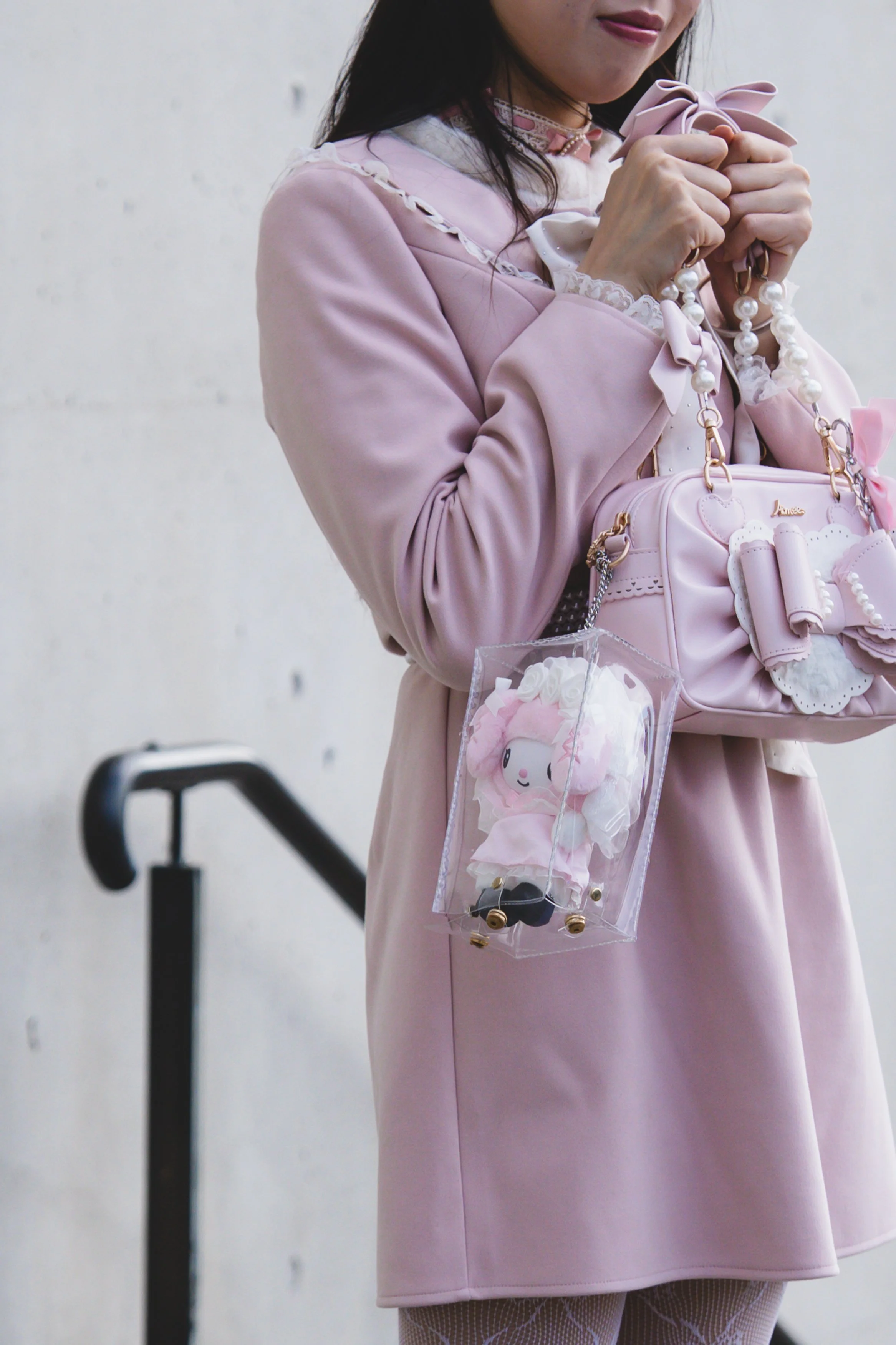 A woman dressed in a pink outfit holding a pink handbag with pearl accents and bows, along with other pastel accessories, standing against a plain light-colored background.