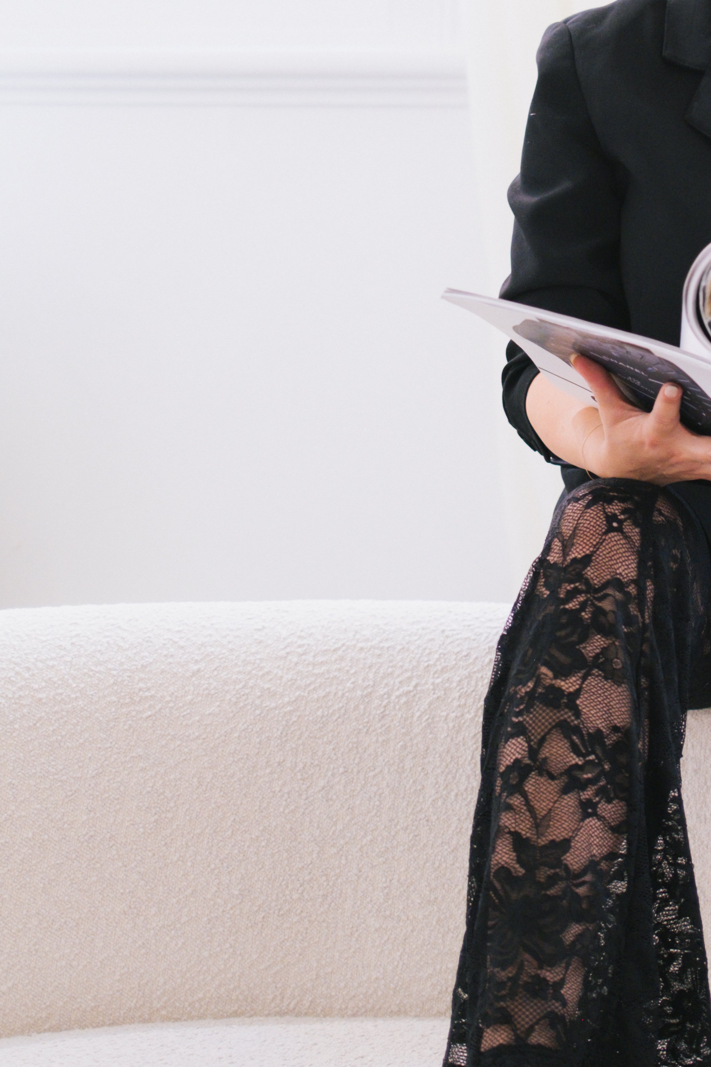 A woman wearing a black blazer, holding magazines, and dressed in black lace stockings, standing in front of a white wall.