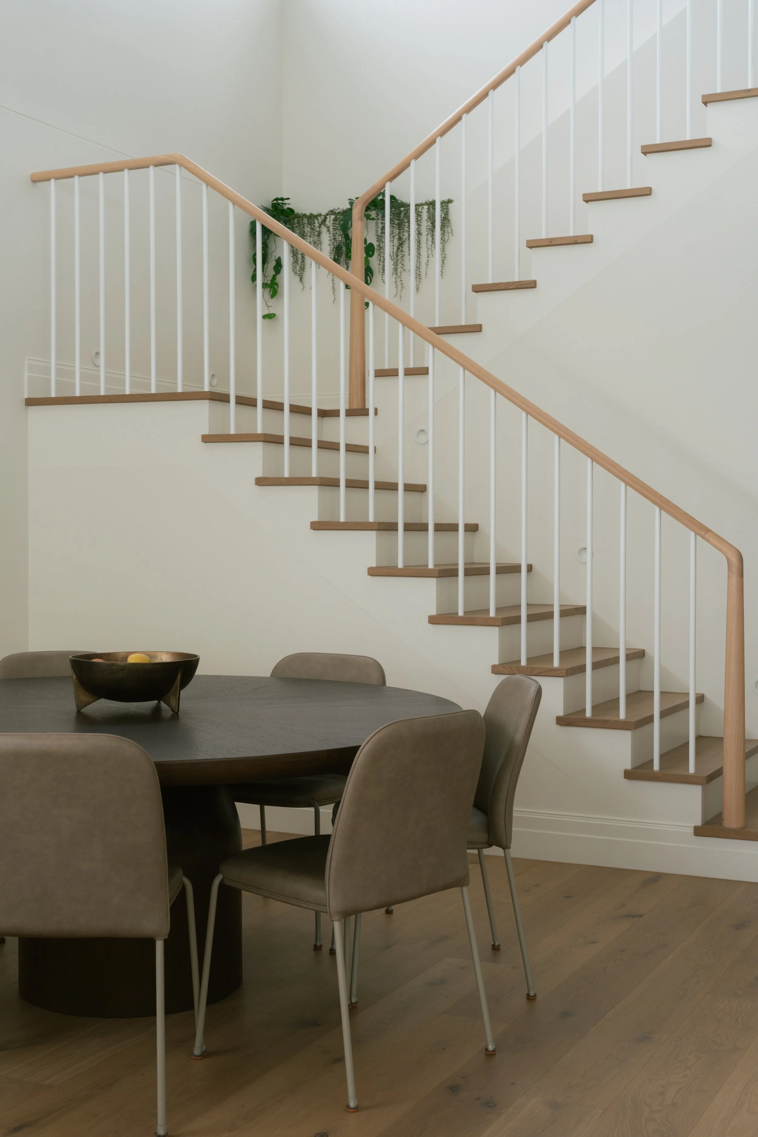 Interior space with a wooden staircase, beige chairs around a dark round dining table, and a bowl with fruit, with a hanging green plant at the top of the stairs.