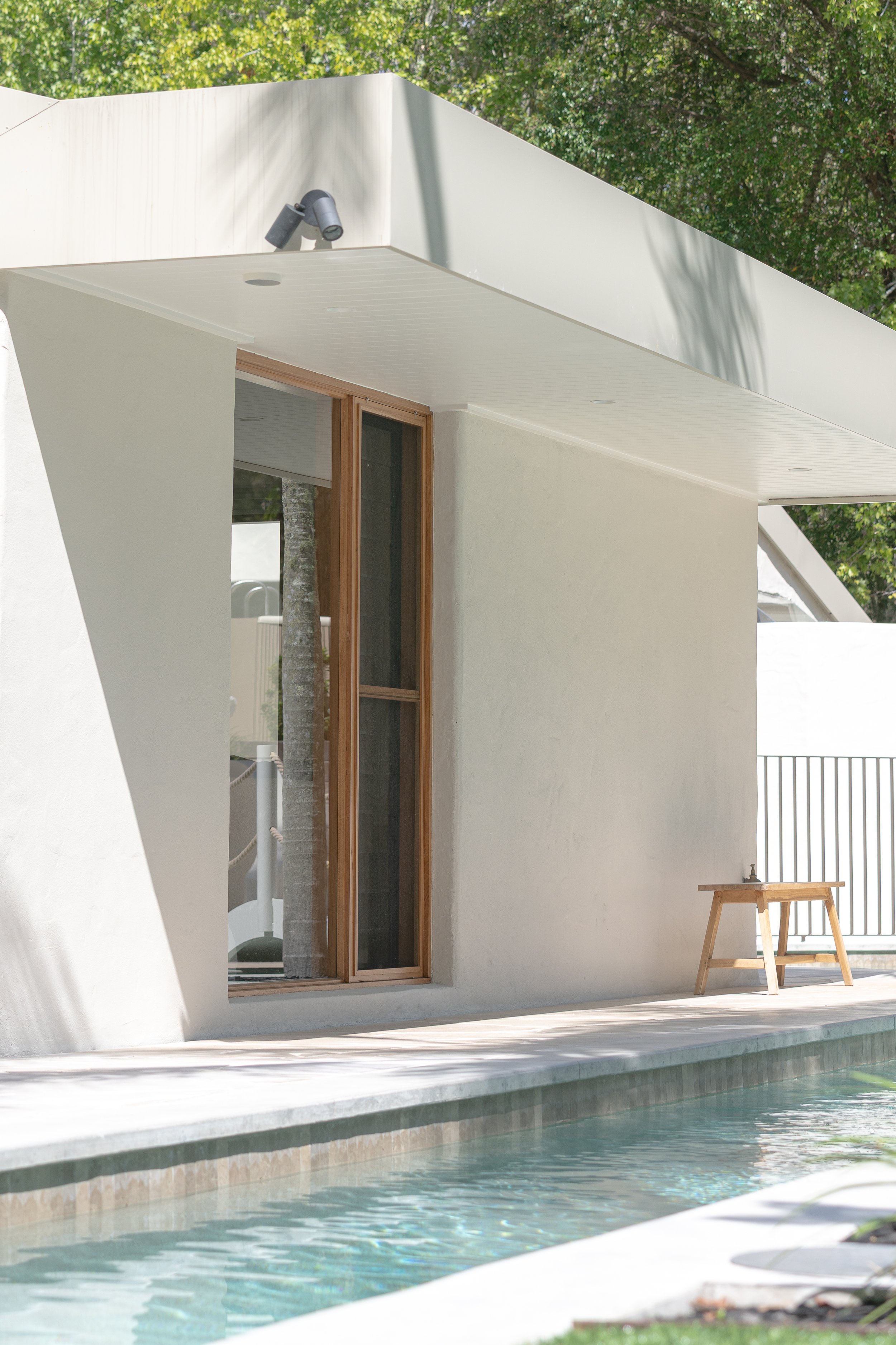 Exterior view of a modern house with a swimming pool, sliding glass door, and a small wooden bench on a patio, surrounded by green trees.