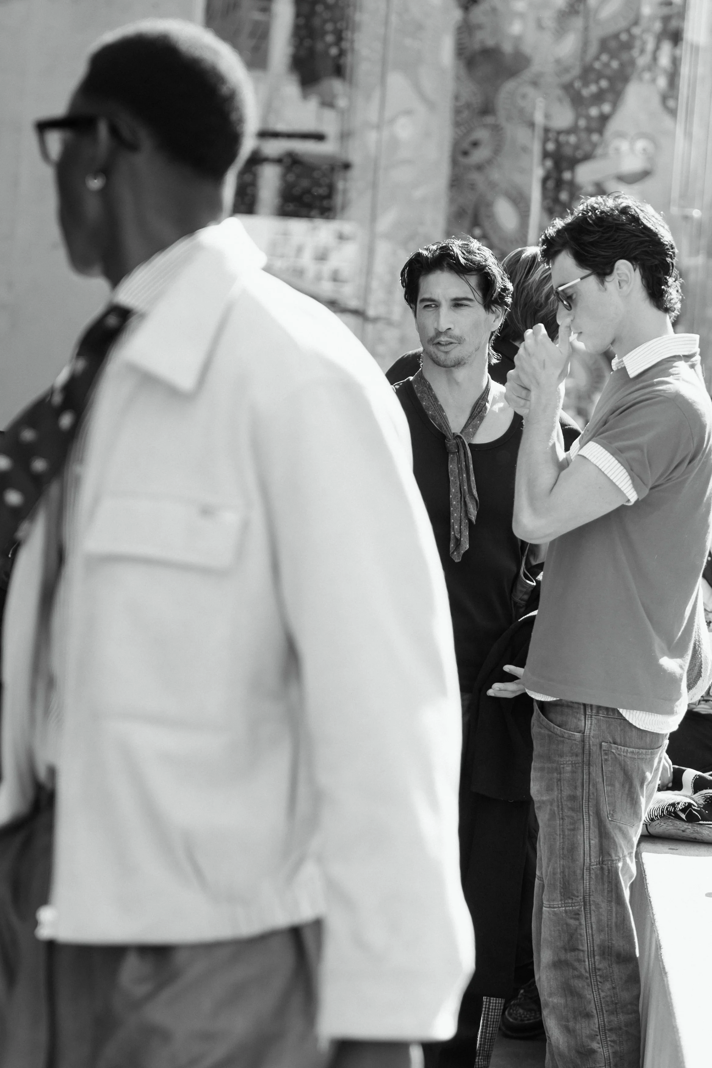 A group of people standing outside on a sunny day, engaged in conversation. The image is in black and white.
