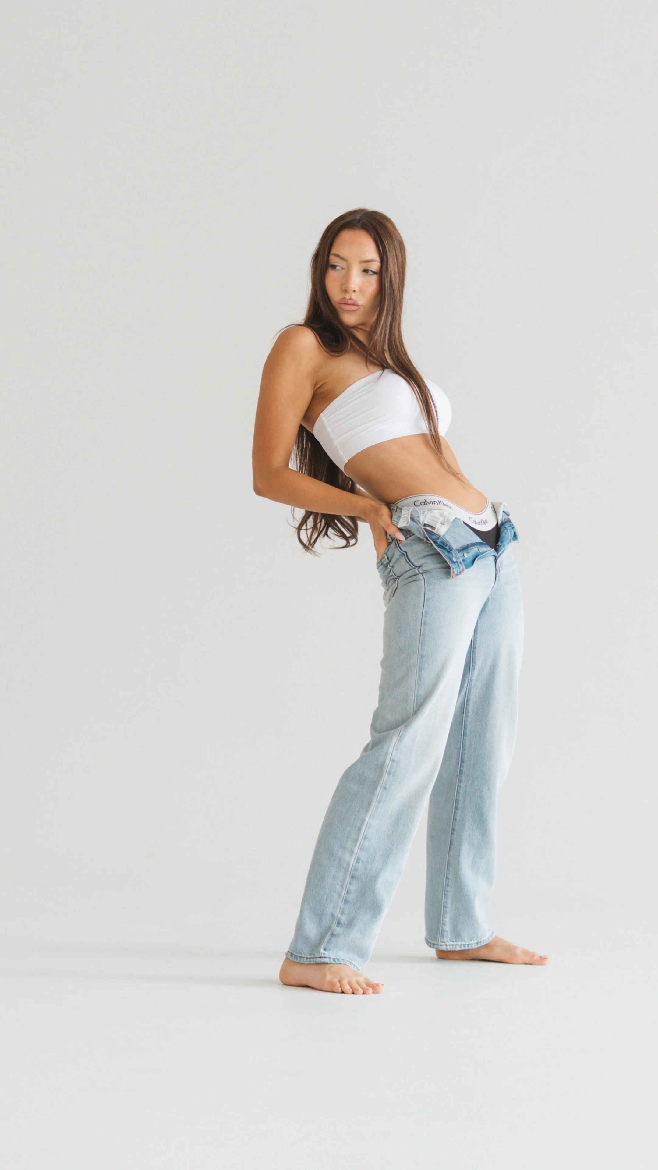 A woman with long brown hair in loose waves standing on a white background wearing a white tube top and light-wash jeans with the waistband slightly rolled down, revealing Calvin Klein underwear. She is barefoot and looking to her left with a relaxed pose.