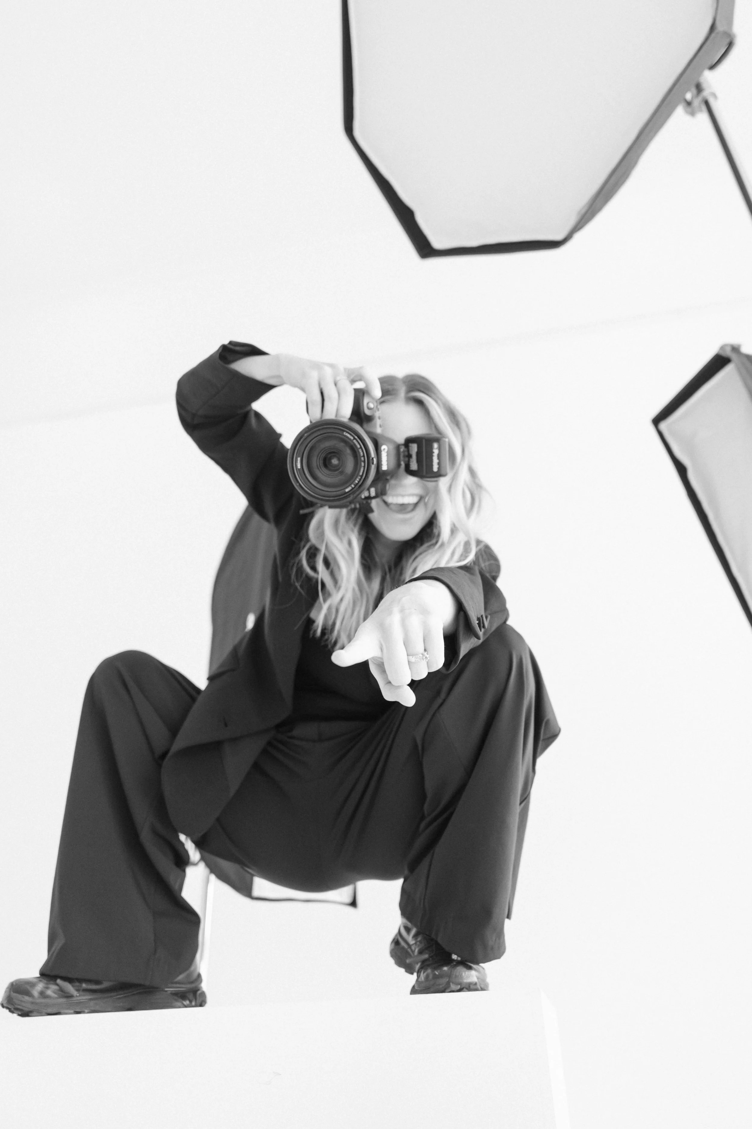 A person dressed in dark clothing crouching down and pointing towards the camera while smiling and holding a camera in front of their face. The background is plain and there are some studio lights visible.