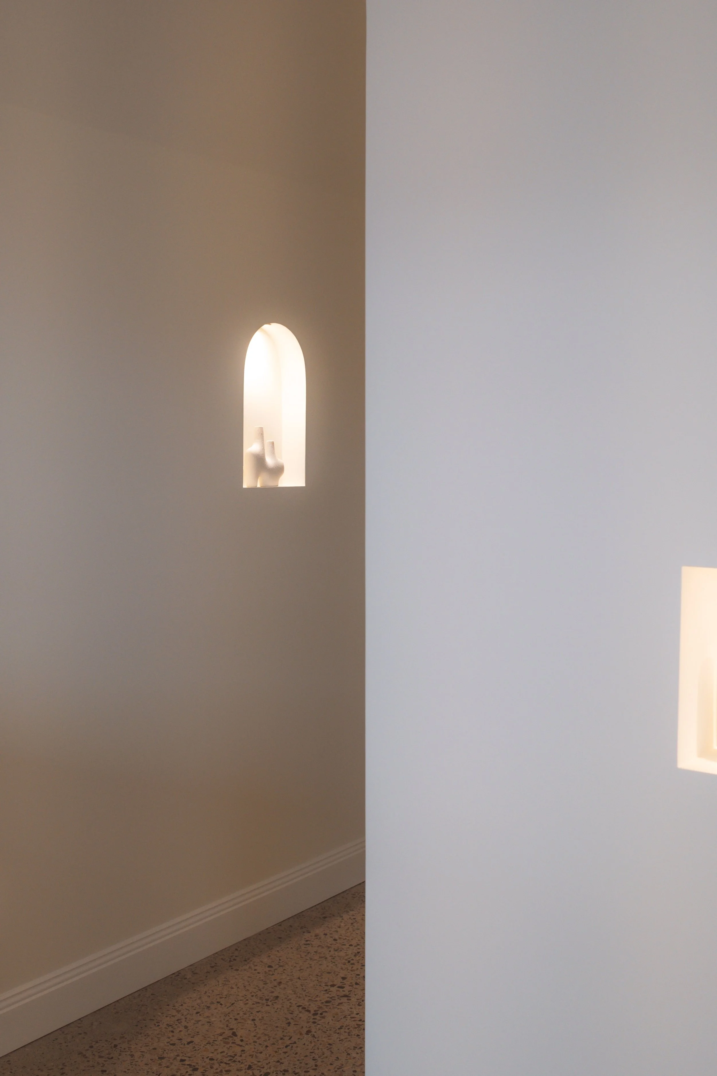 A hallway with beige walls and a speckled floor, partially obscured by a white wall or door in the foreground. There is a small, arched recess in the beige wall, containing a white sculpture of two figures, illuminated by a light.