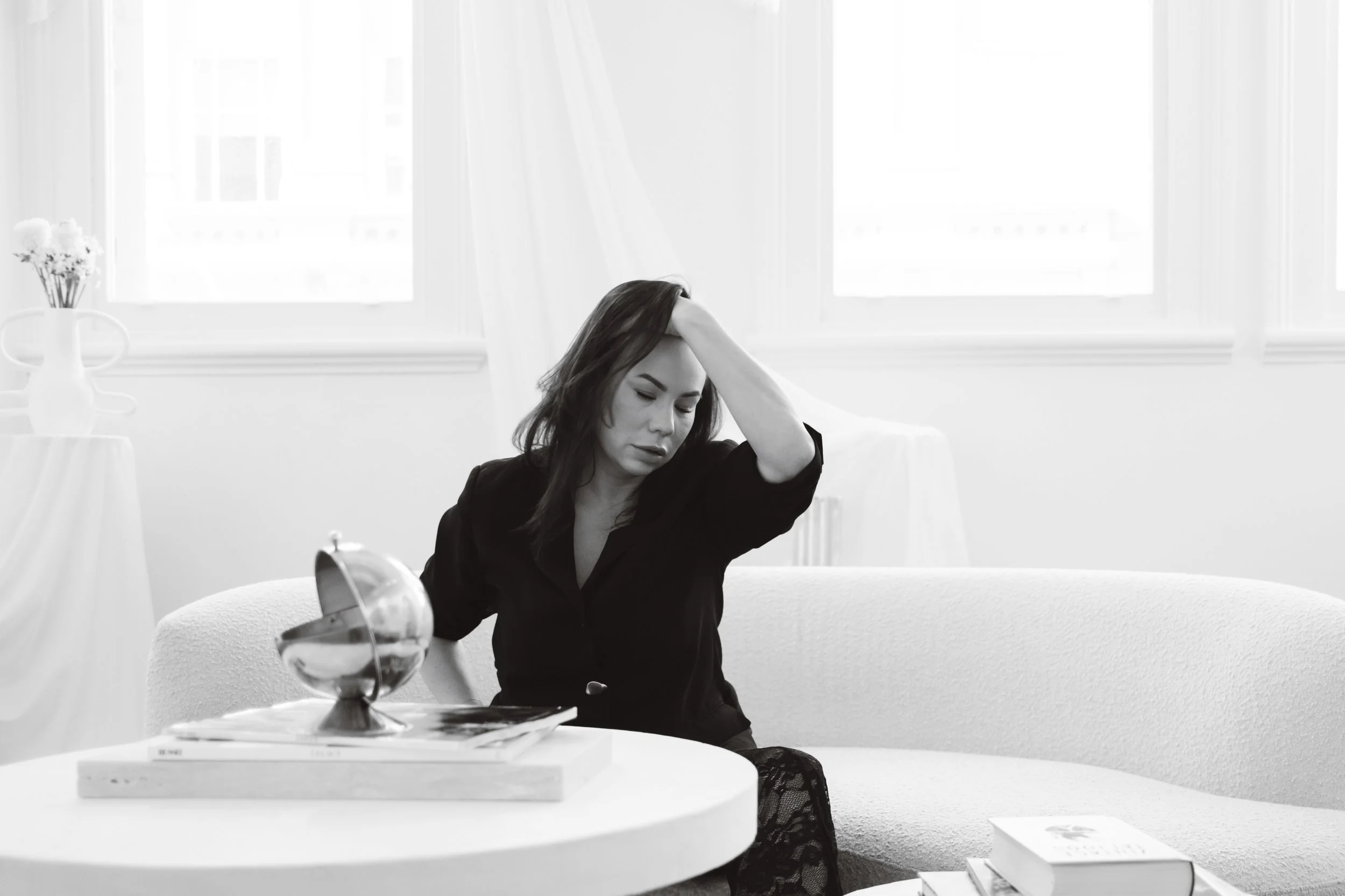 A woman sitting on a couch with her hand on her head, facing downward. A table in front with a mirror, books, and a lamp. Bright windows in the background.