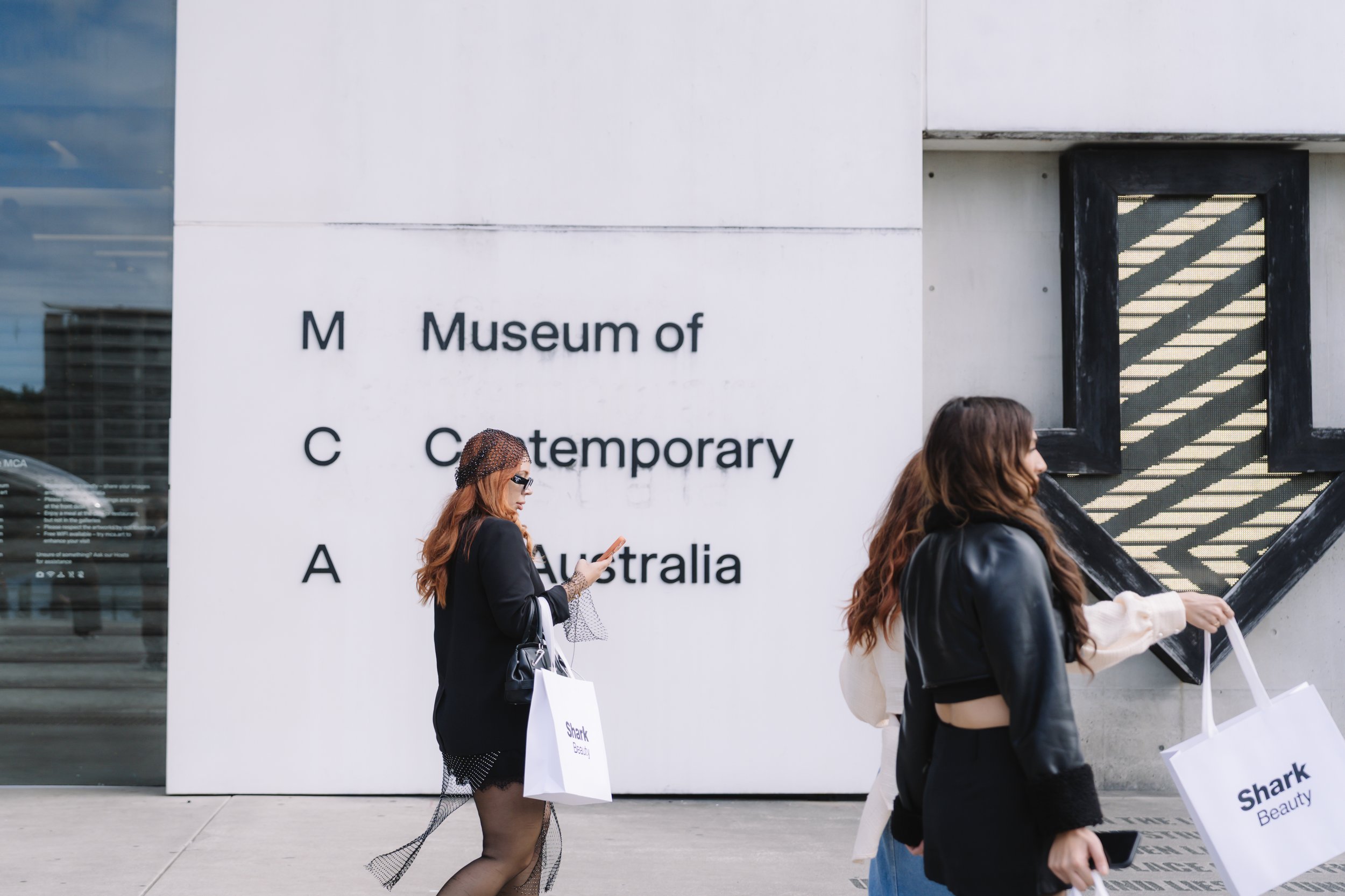 People walking past the Museum of Contemporary Australia building, with signage and shadows on the wall.