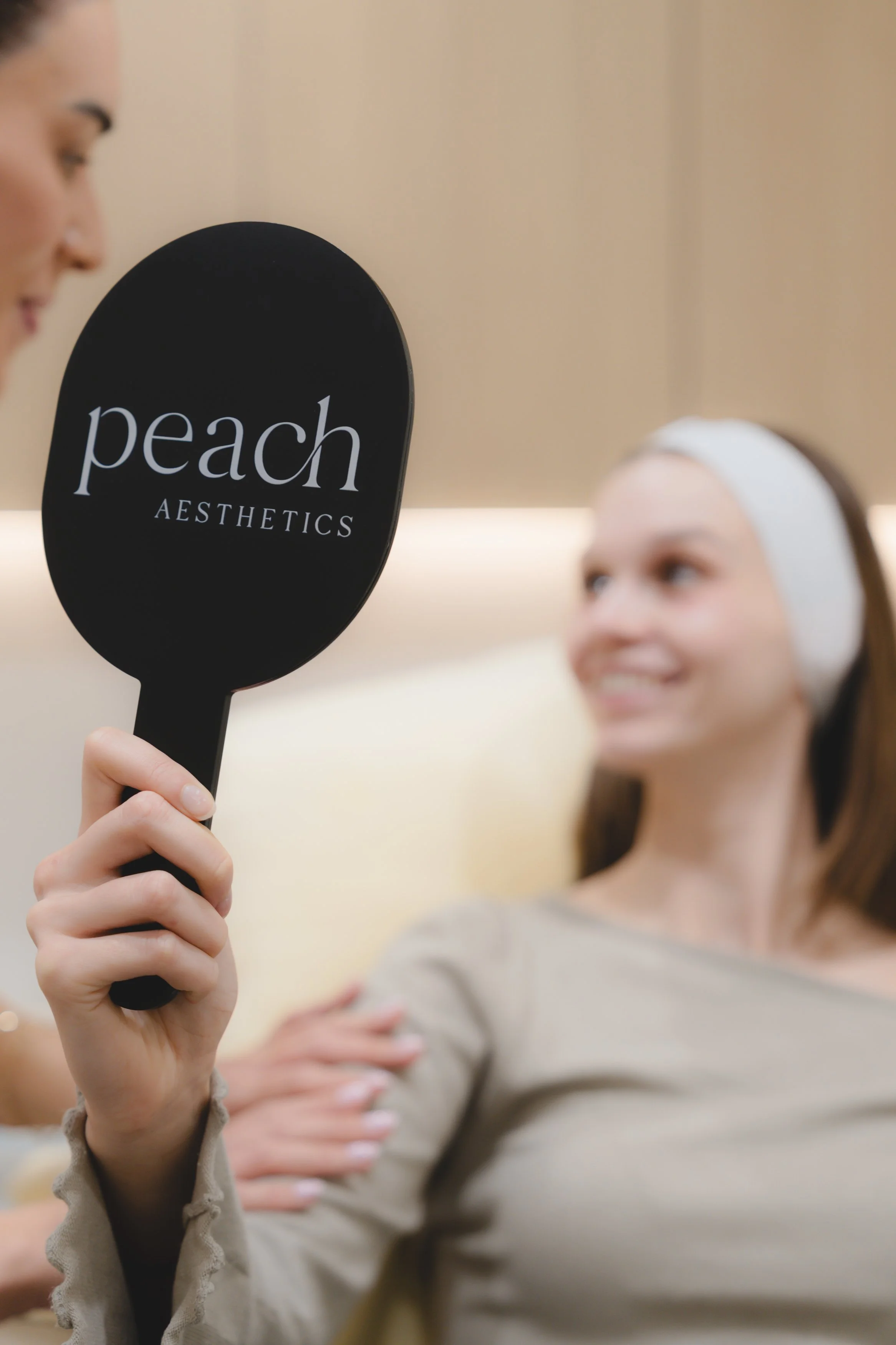 A woman holding a hand mirror with a black handle and black back that reads 'peach AESTHETICS'; she is smiling and looking at her reflection, with her arms crossed.