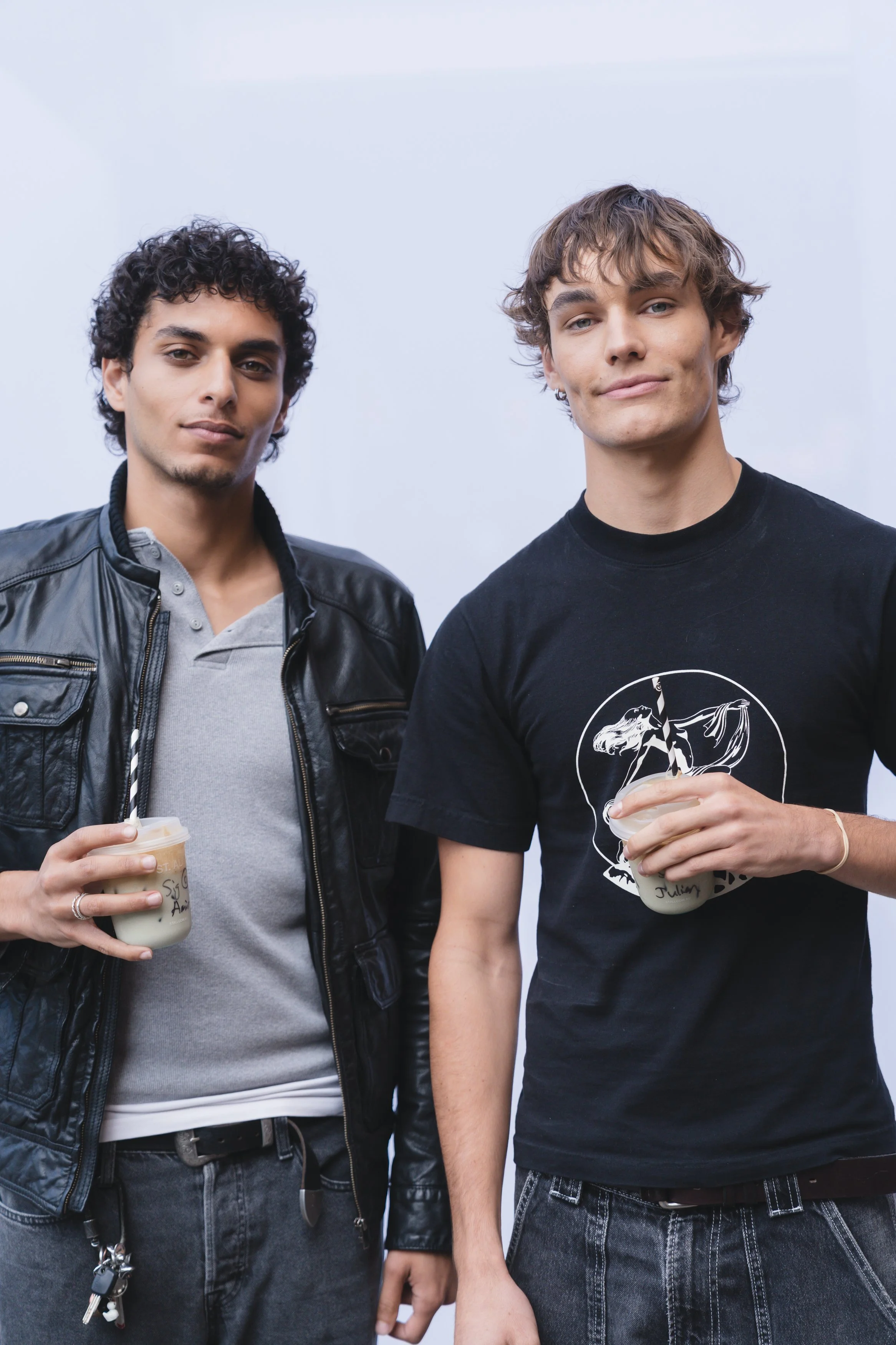 Two young men with light skin holding beverages, standing close together, and looking at the camera. One has curly dark hair and is wearing a black leather jacket, while the other has wavy brown hair and is wearing a black T-shirt with a graphic.