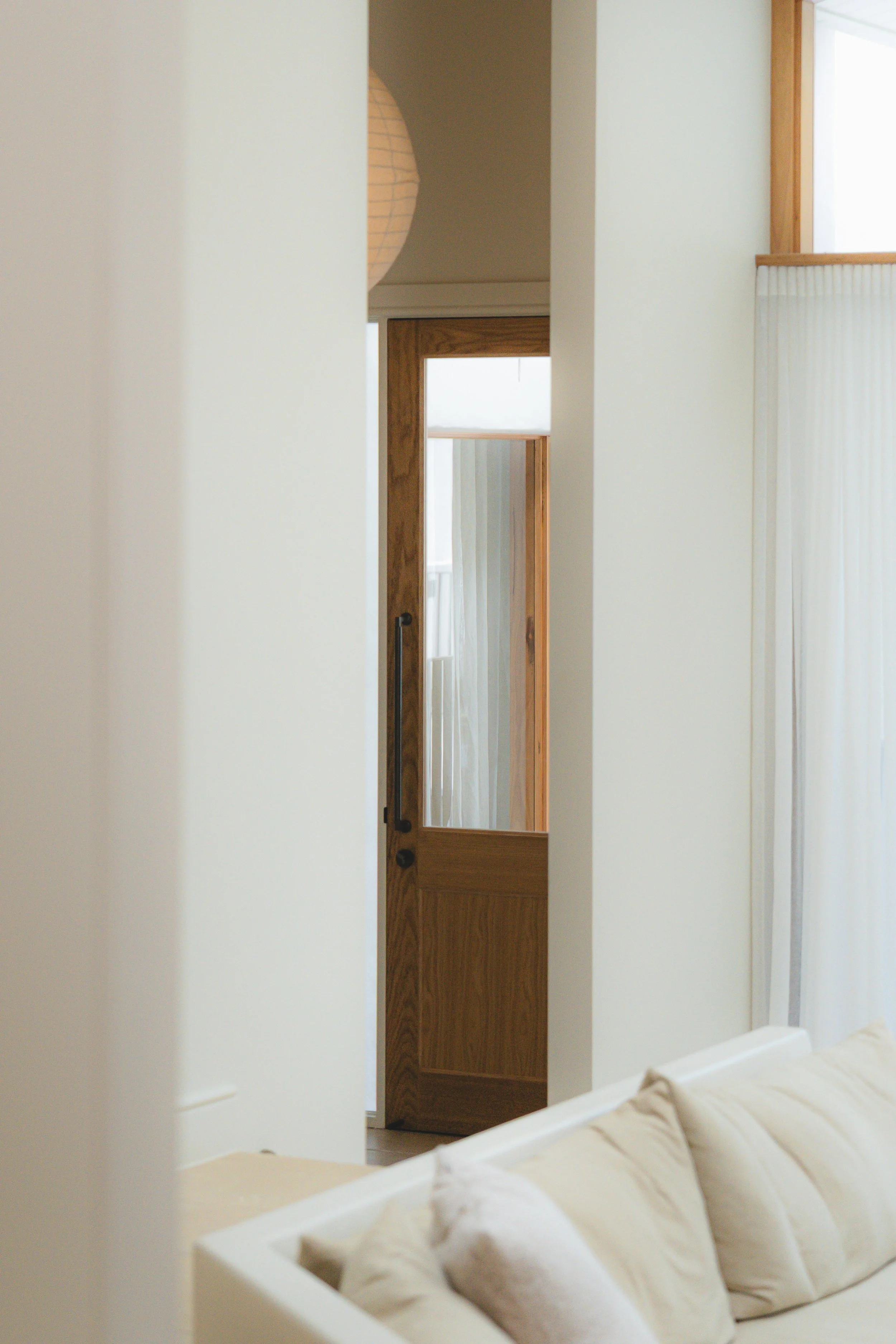 A bedroom with a wooden door and white walls, partially visible beige sofa with pillows, and a white decorative basket on the bed.