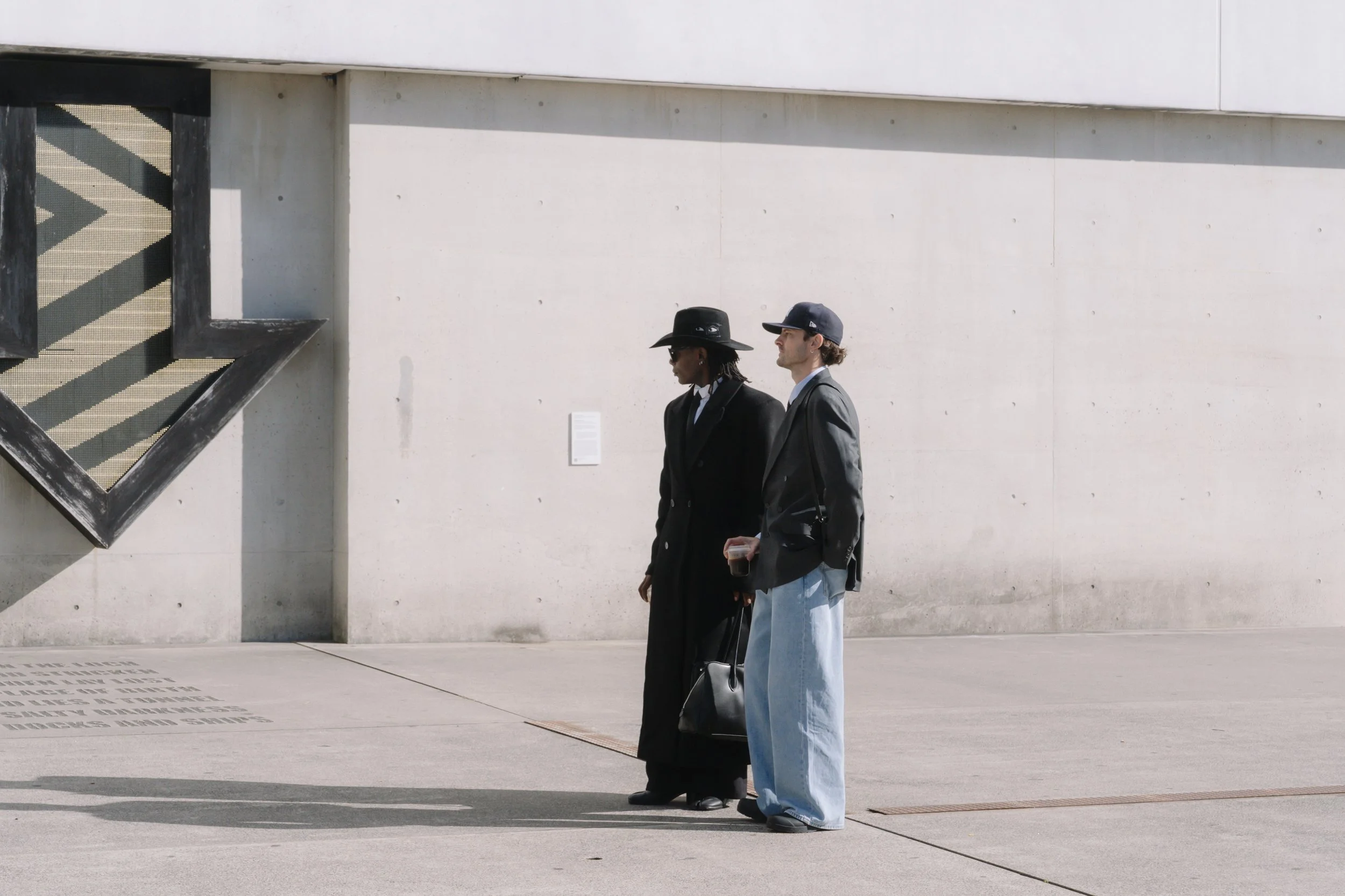 Two people walking side by side in front of a white wall. The person on the left is wearing a black wide-brimmed hat, black coat, and sunglasses. The person on the right is wearing a gray cap, gray blazer, and light blue wide-leg jeans, holding a drink in their right hand. There is minimal shadow cast on the wall and ground.