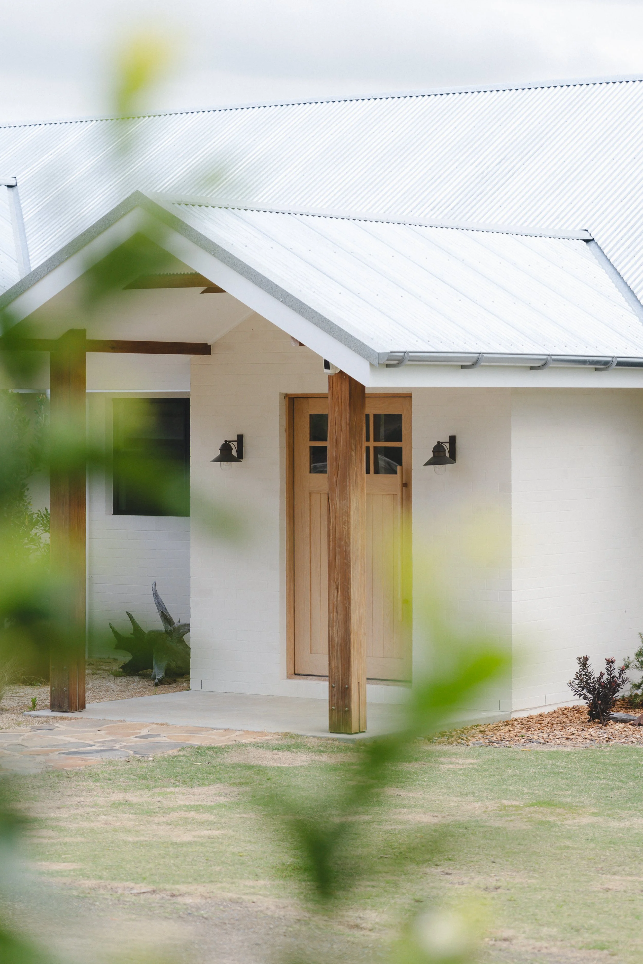 The image shows the exterior of a modern house with a white brick wall, large wooden door, and two wall-mounted outdoor lights. The roof has a sloped metal design with a gutter, supported by wooden posts, and is partially obscured by some greenery in