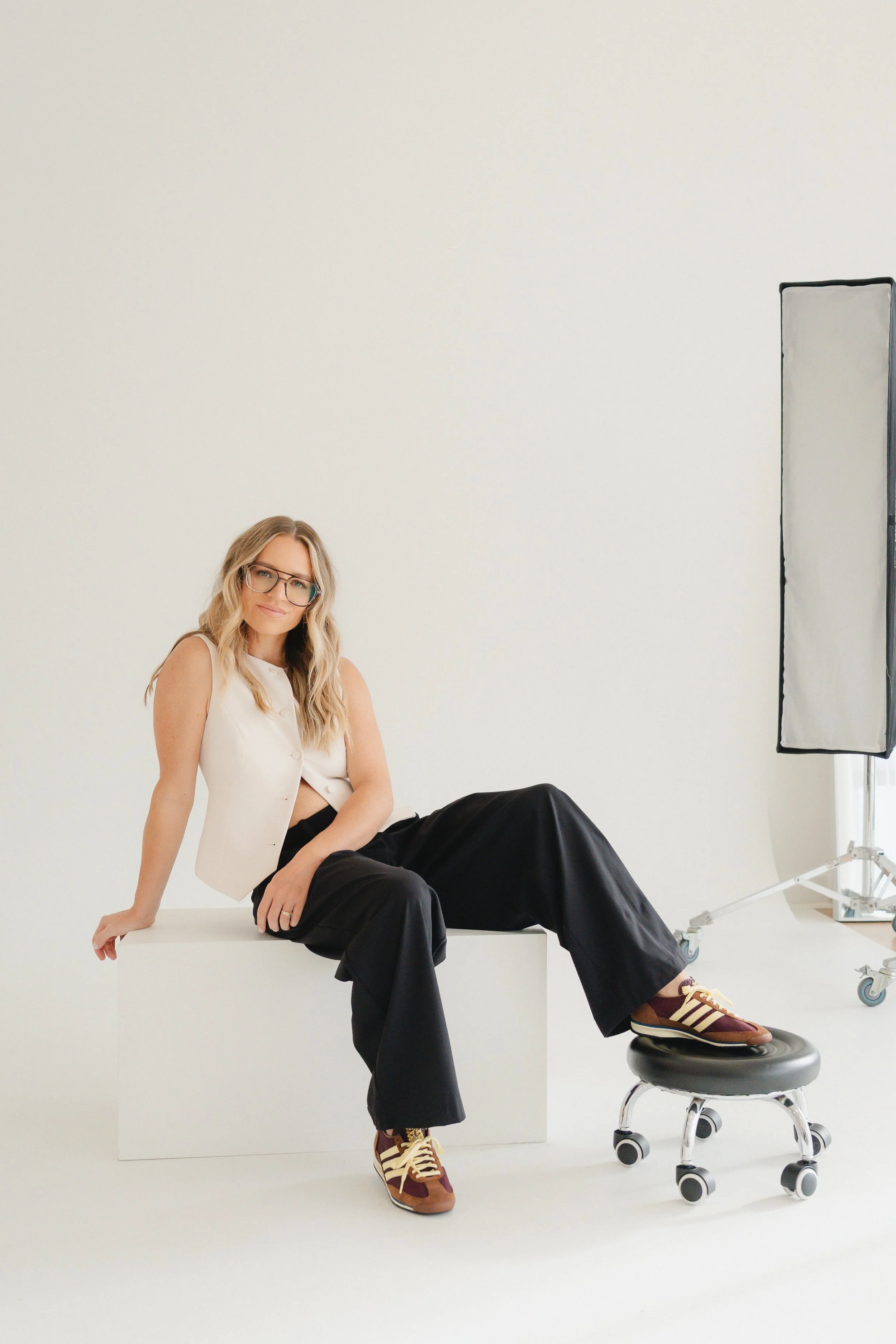 A woman with blonde hair, glasses, and a white sleeveless top sitting on a white box, with one leg bent and resting on an examination table, and the other leg extended down, wearing black pants and brown sneakers with white stripes. She is smiling an