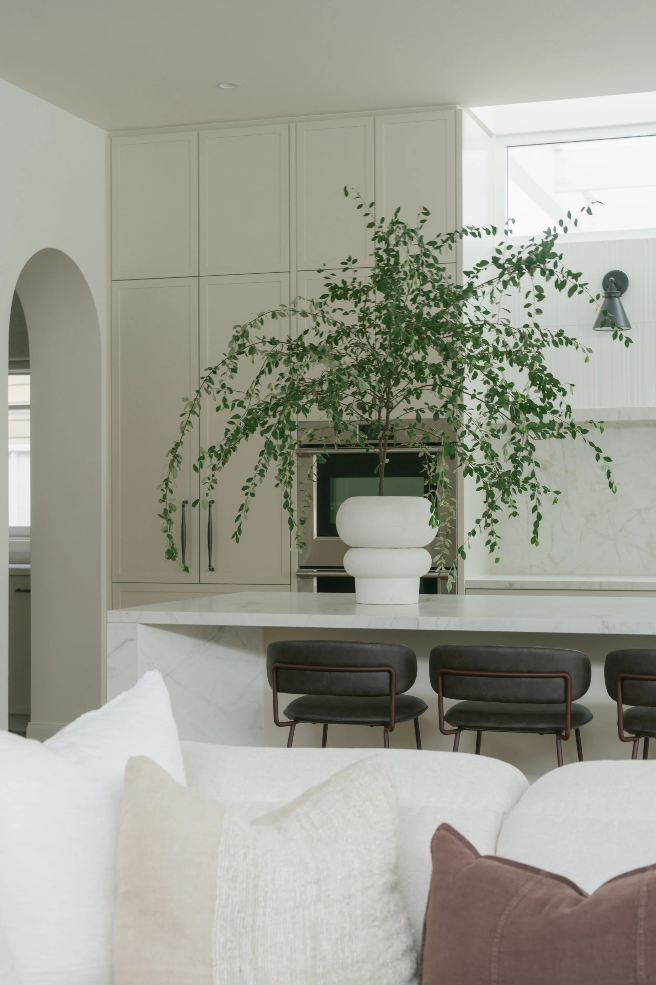 A modern kitchen with white walls, cabinetry, and a marble kitchen island. A large potted plant sits in the center of the island. Black chairs are positioned at the kitchen counter, and natural light streams in from a window near the ceiling.