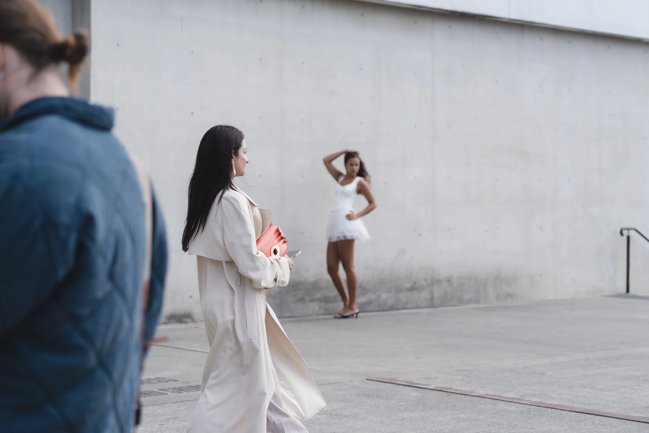A woman in a beige coat walks past a woman in a white dress posing against a concrete wall. The woman in the white dress has her left hand on her hip and her right arm raised, with her hand behind her head. A person with brown hair and a ponytail, wearing glasses and a denim jacket, is partly visible on the left.