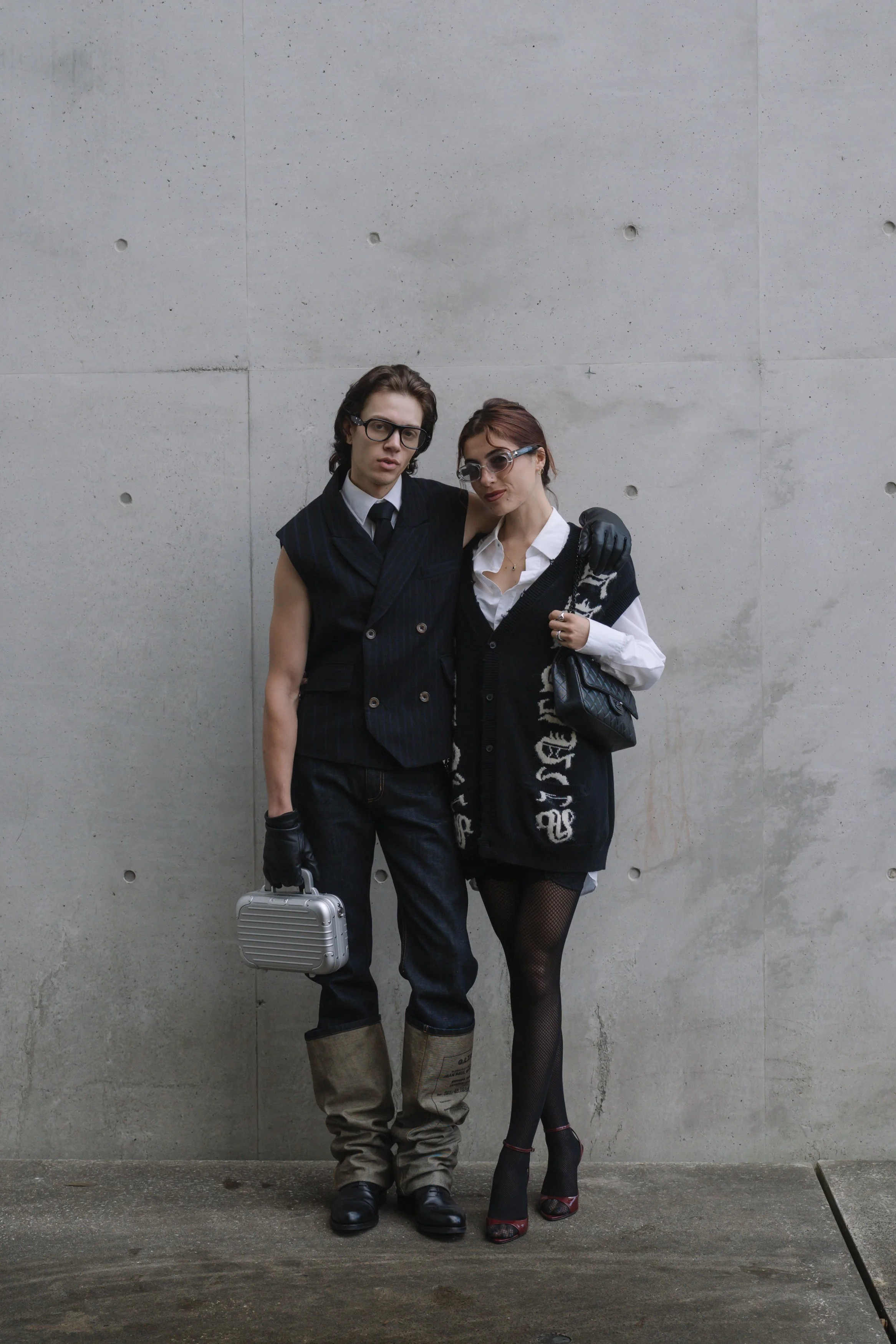 Two individuals, one man and one woman, standing close together against a plain concrete wall. The man is dressed in a dark pinstripe vest with a white shirt, black tie, and beige boots, holding a silver briefcase, wearing glasses with a serious expression. The woman is dressed in a black and white sweater with text, a white shirt, fishnet stockings, and high heels, with her arm around the man's shoulders, wearing sunglasses and a slight smile.