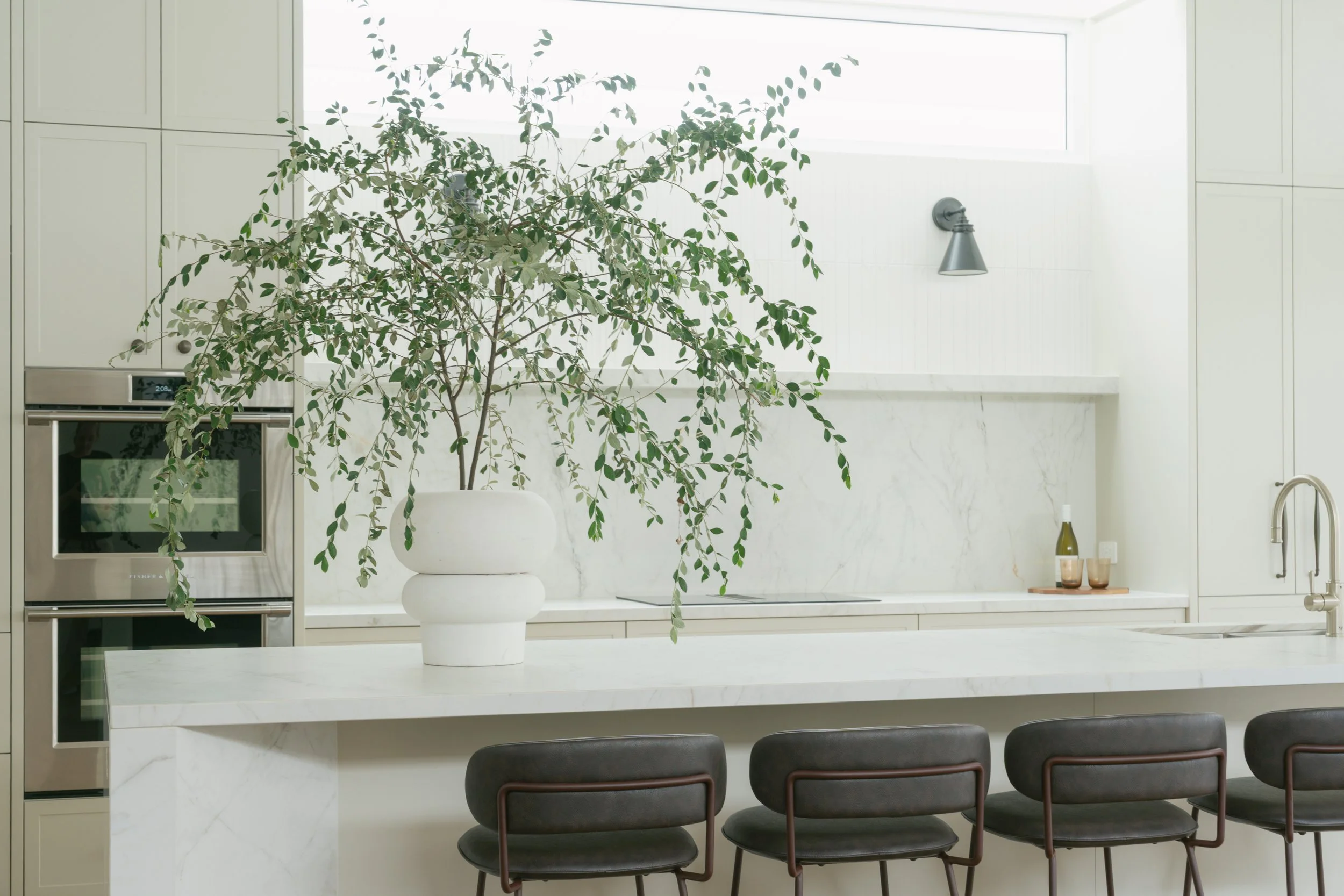 A modern kitchen with white marble countertops and cabinetry, a large potted green plant on the island, and three black chairs in the foreground.
