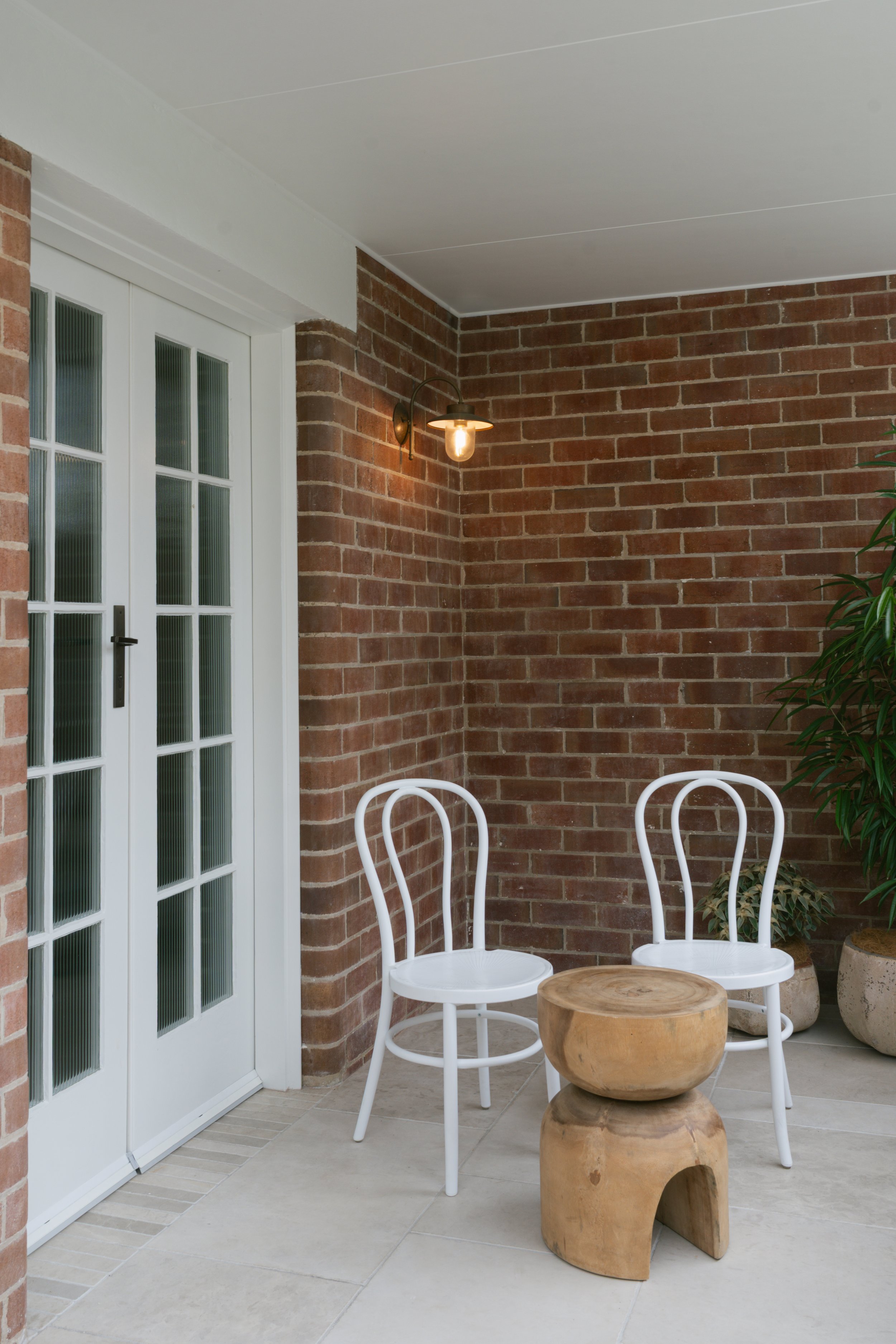 A cozy outdoor patio area with brick walls, a white door with glass panes, a wall-mounted vintage-style light fixture, three white chairs, a small wooden stool, and potted plants.